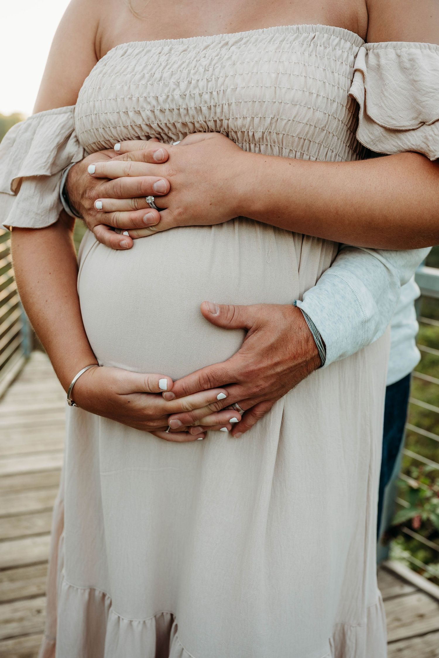 A pregnant woman in a flowy beige dress holds her belly while loving hands embrace her from behind on a wooden deck.