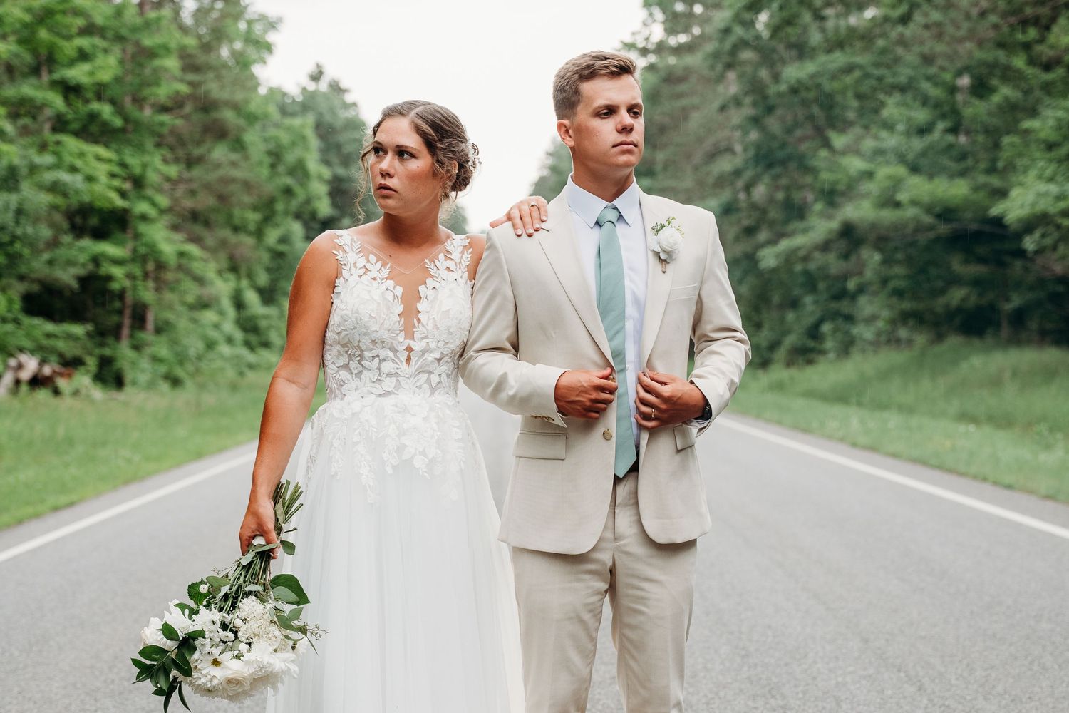 A couple in wedding attire walks down a winding country road surrounded by lush green trees and foliage.