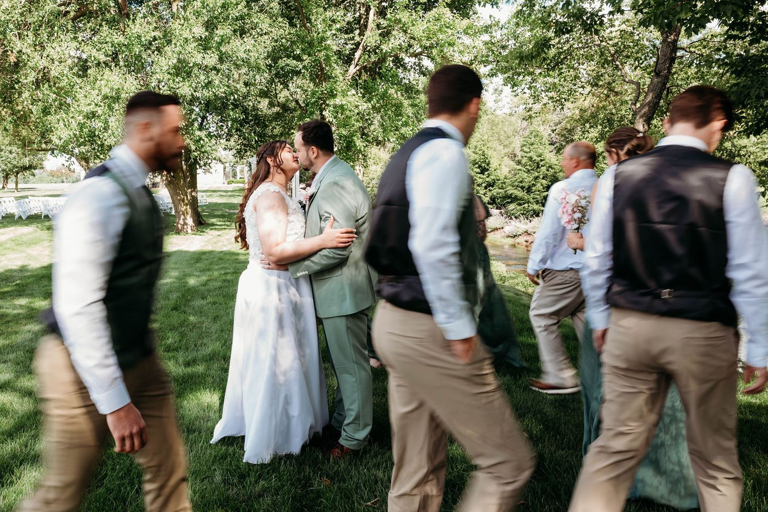 Bride and groom share a kiss outdoors at their wedding reception surrounded by groomsmen in vests and khakis.