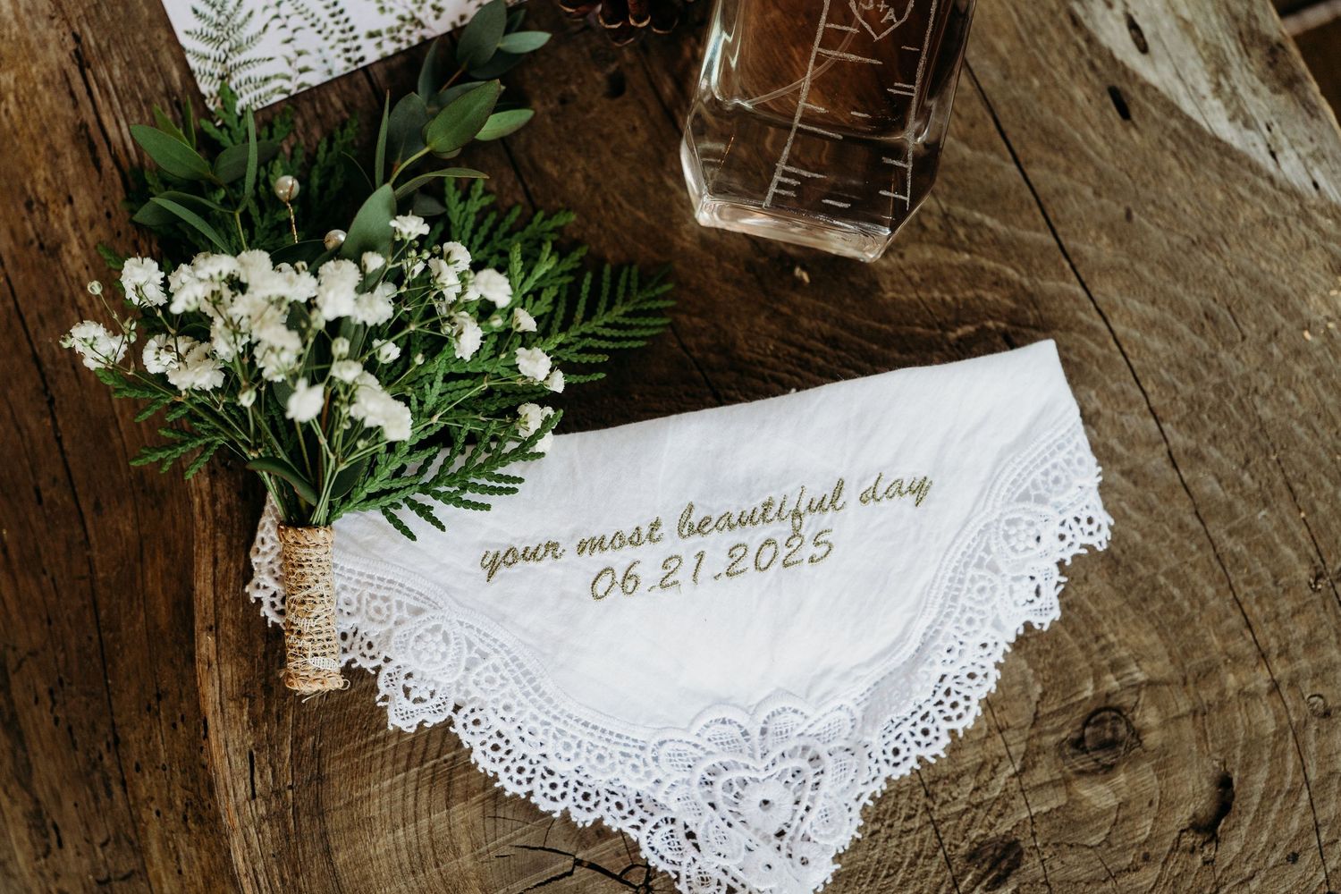 White lace handkerchief with embroidered date and small floral arrangement on wooden surface.