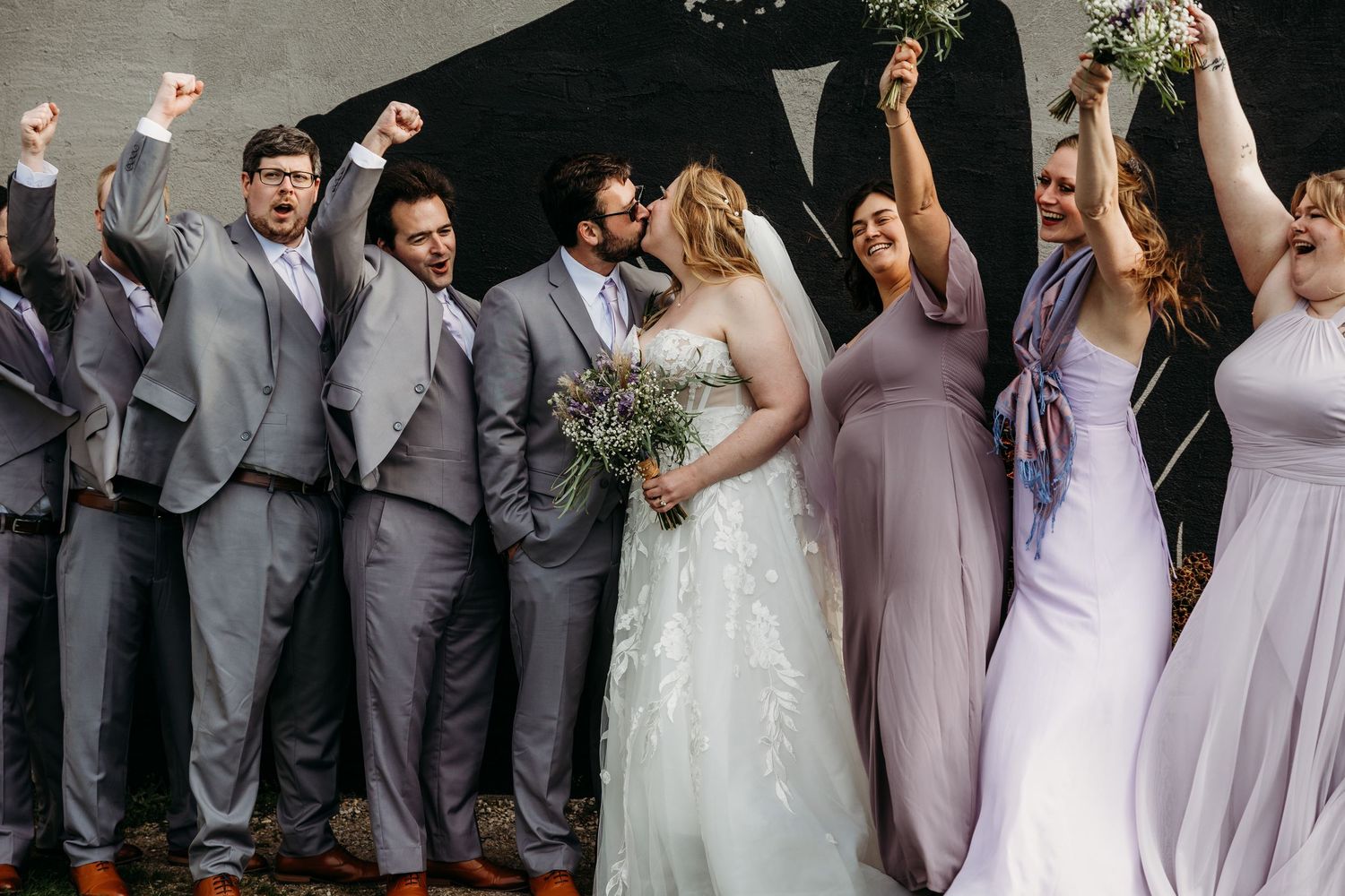 Wedding party members in gray suits and lavender dresses celebrate joyfully in a candid group photo.