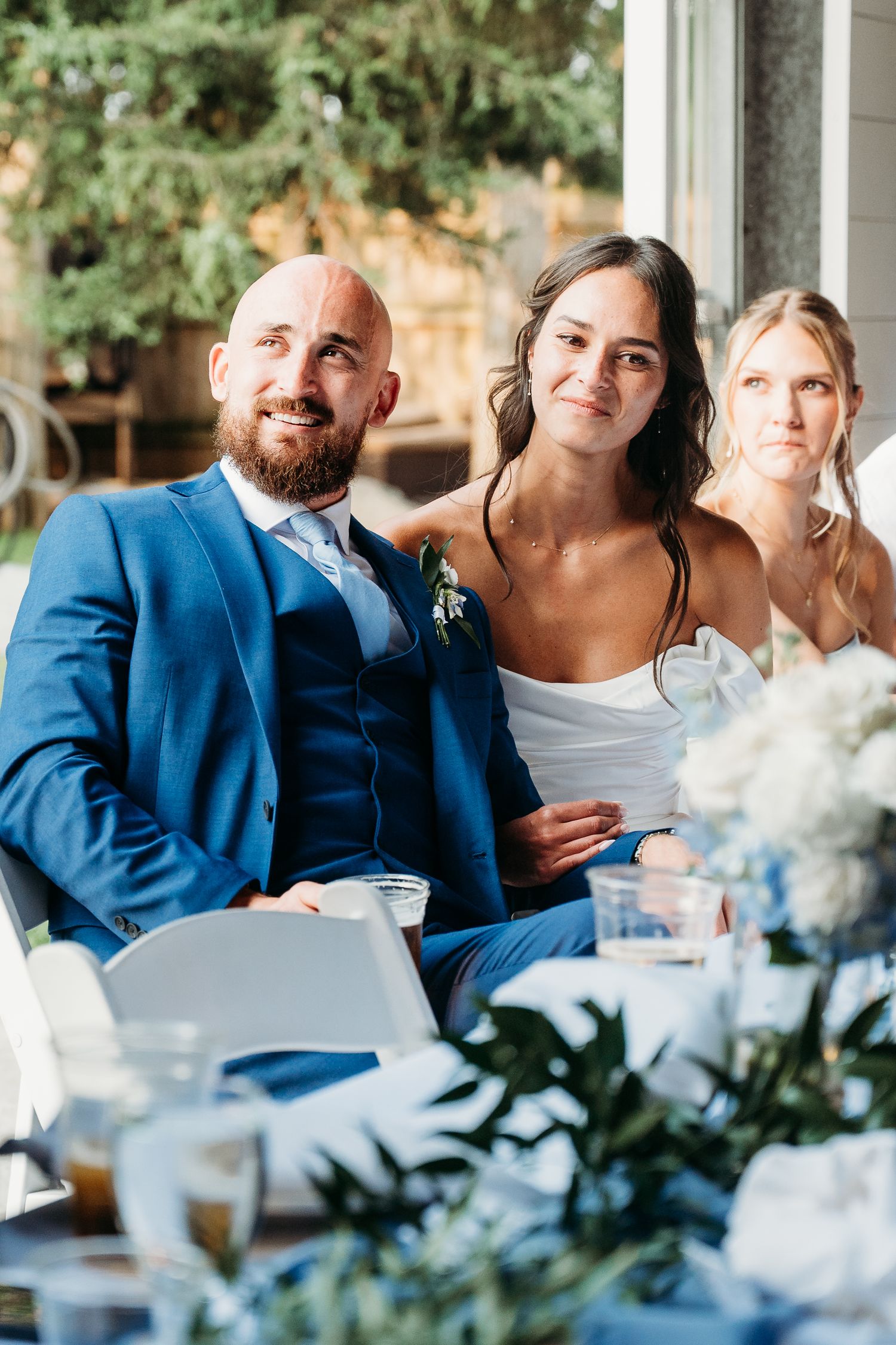 Groom in blue suit and bride in white dress seated at wedding reception table with guests behind them.