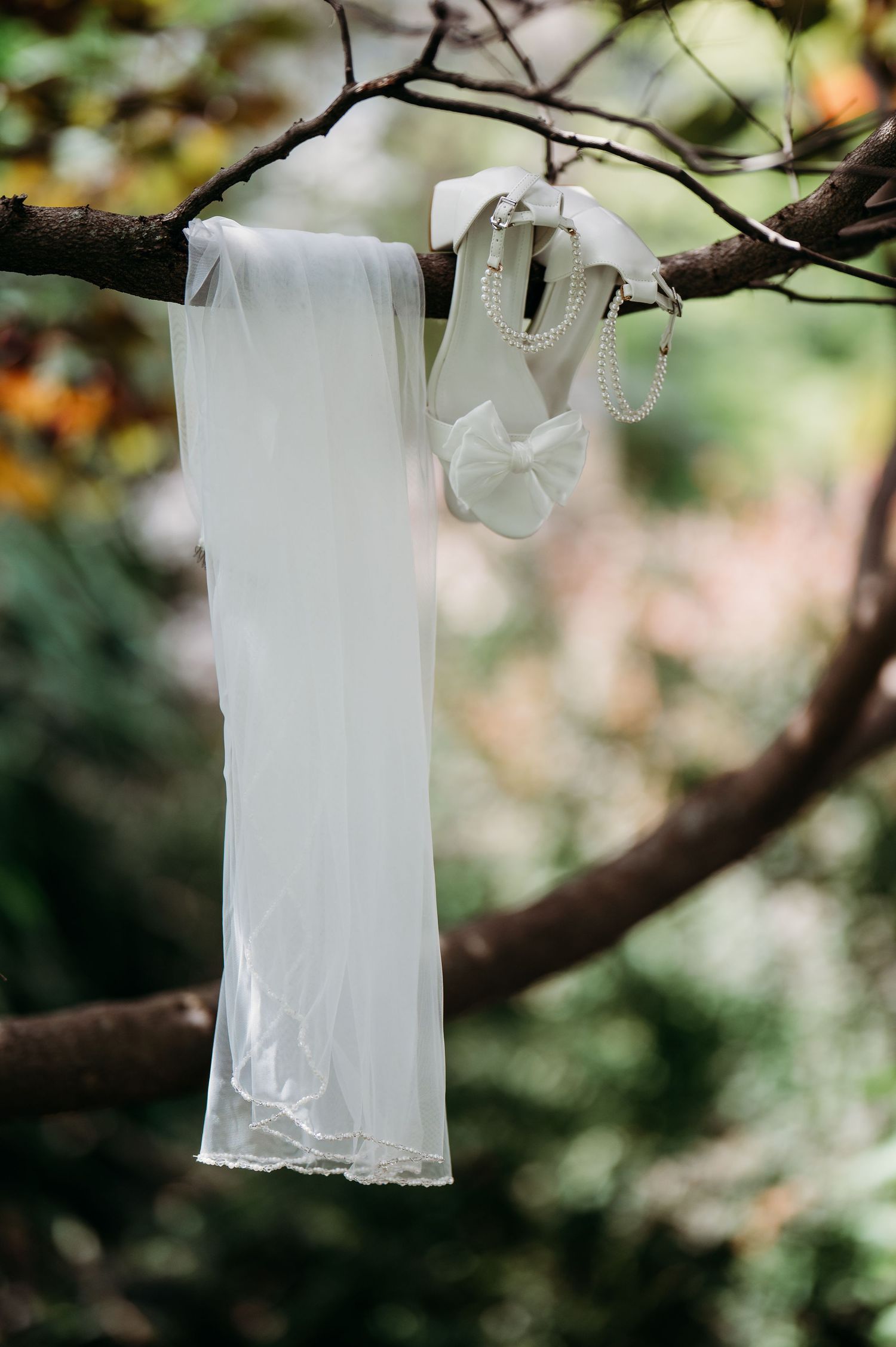 White wedding veil and pearl bridal accessories hanging elegantly from a tree branch in a lush garden setting.