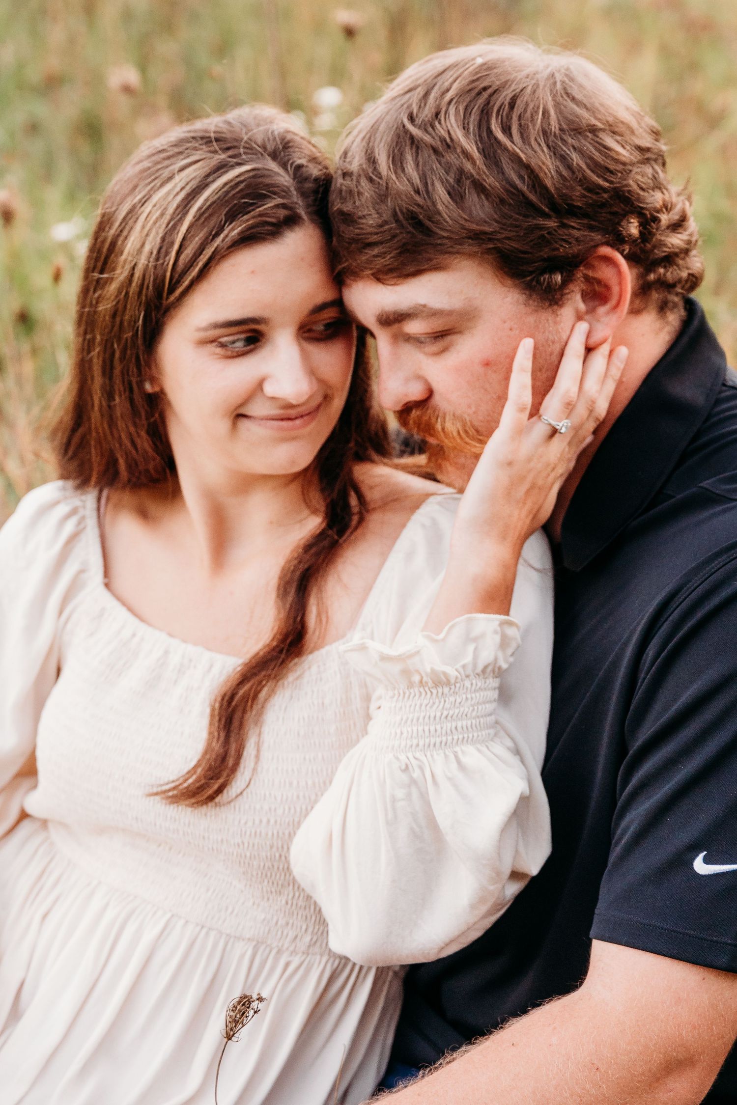 A couple shares an intimate moment outdoors, with the woman in a white dress touching her partner's face tenderly.