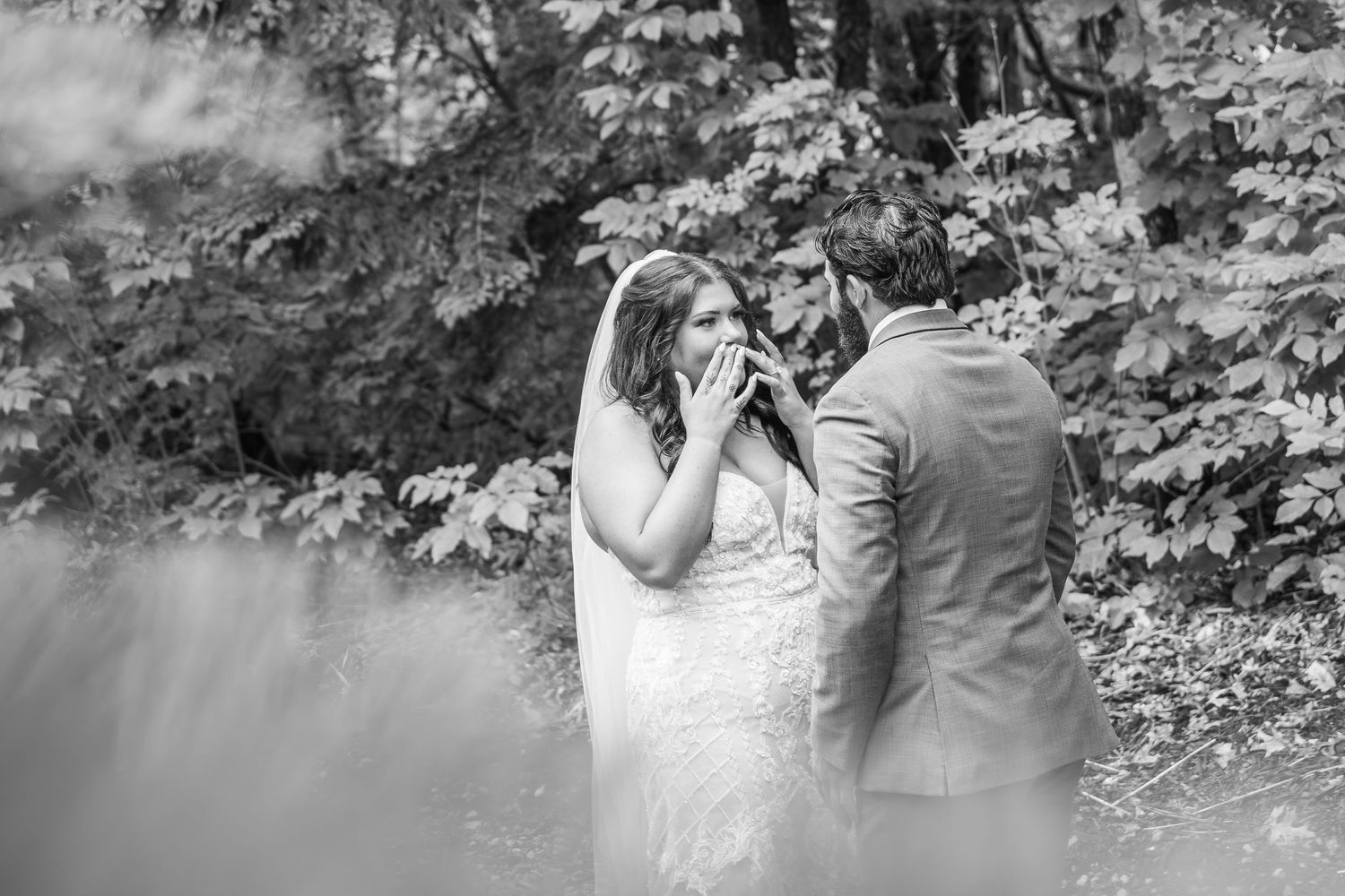 Black and white photo of emotional bride covering her mouth while groom holds her hands outdoors among lush trees.