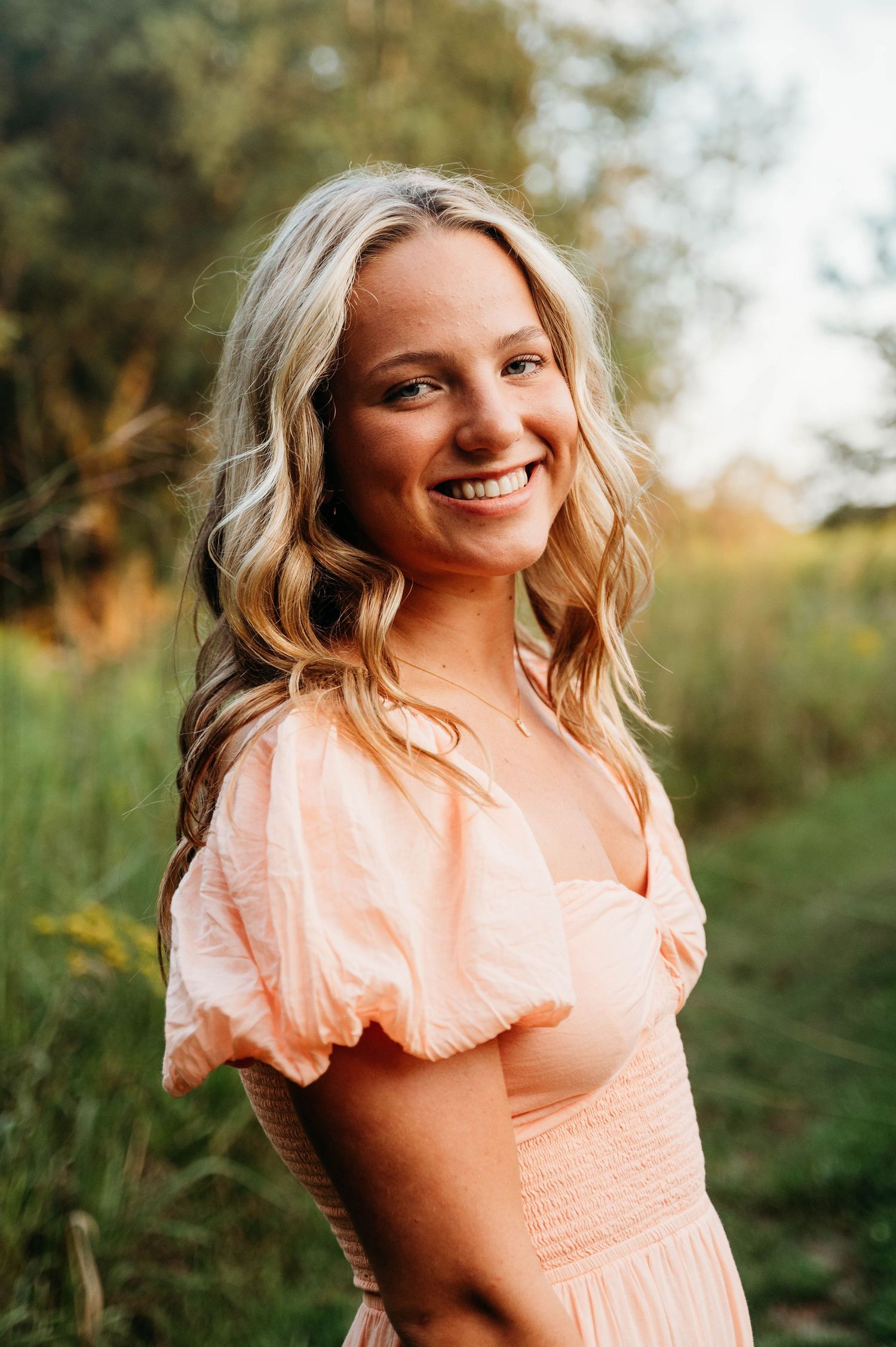 A person wearing a peach-colored dress smiles warmly in a natural outdoor setting with soft golden lighting.