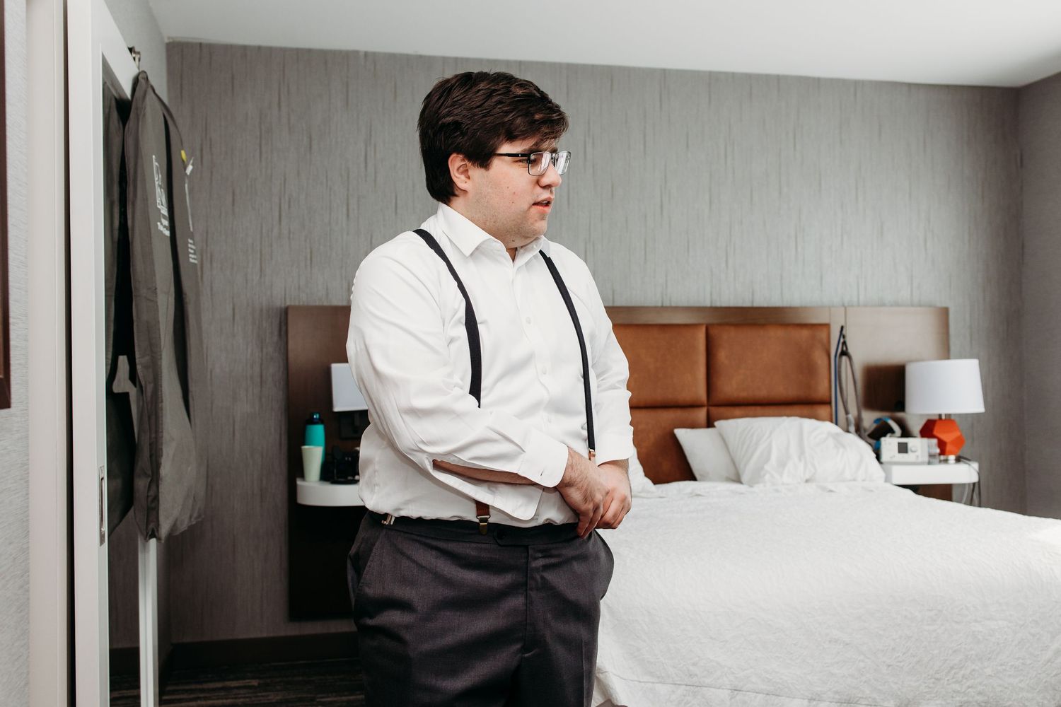 A person in suspenders and white dress shirt standing by a bed in a hotel room.