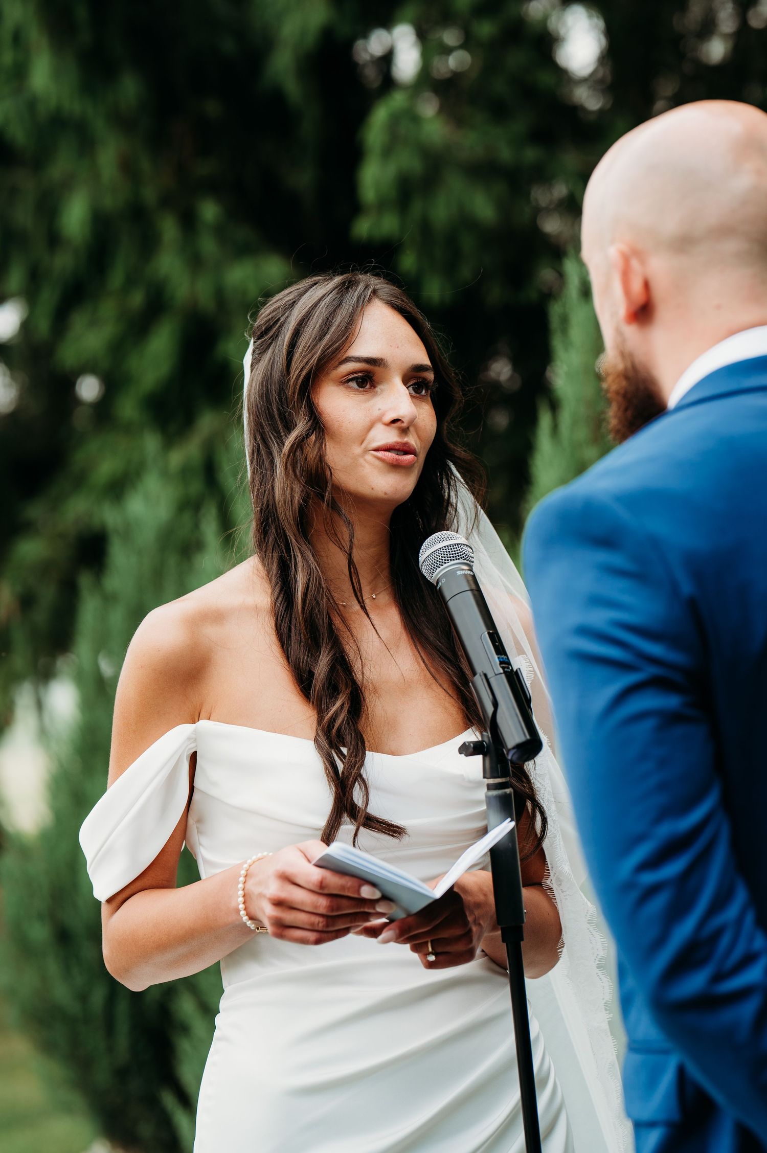 A couple exchanges vows during an outdoor garden wedding ceremony beneath evergreen trees.