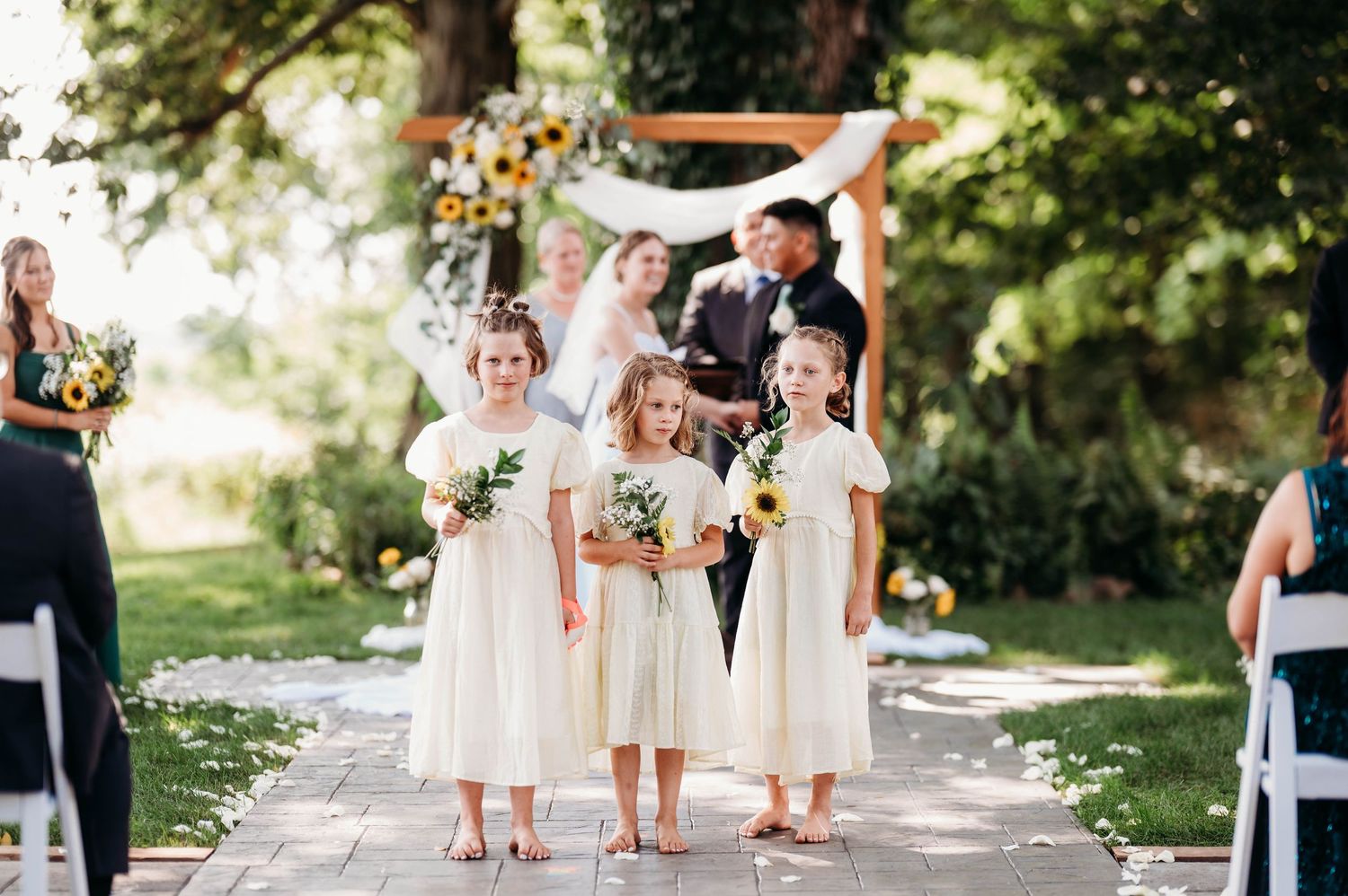 Three flower girls in white dresses walk down outdoor wedding aisle with sunflower bouquets during ceremony.
