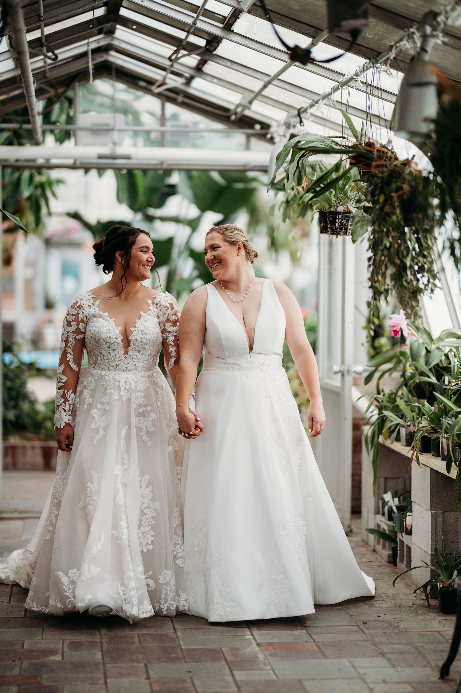 Wedding couple in white lace gowns pose together in a greenhouse setting with natural light streaming through.