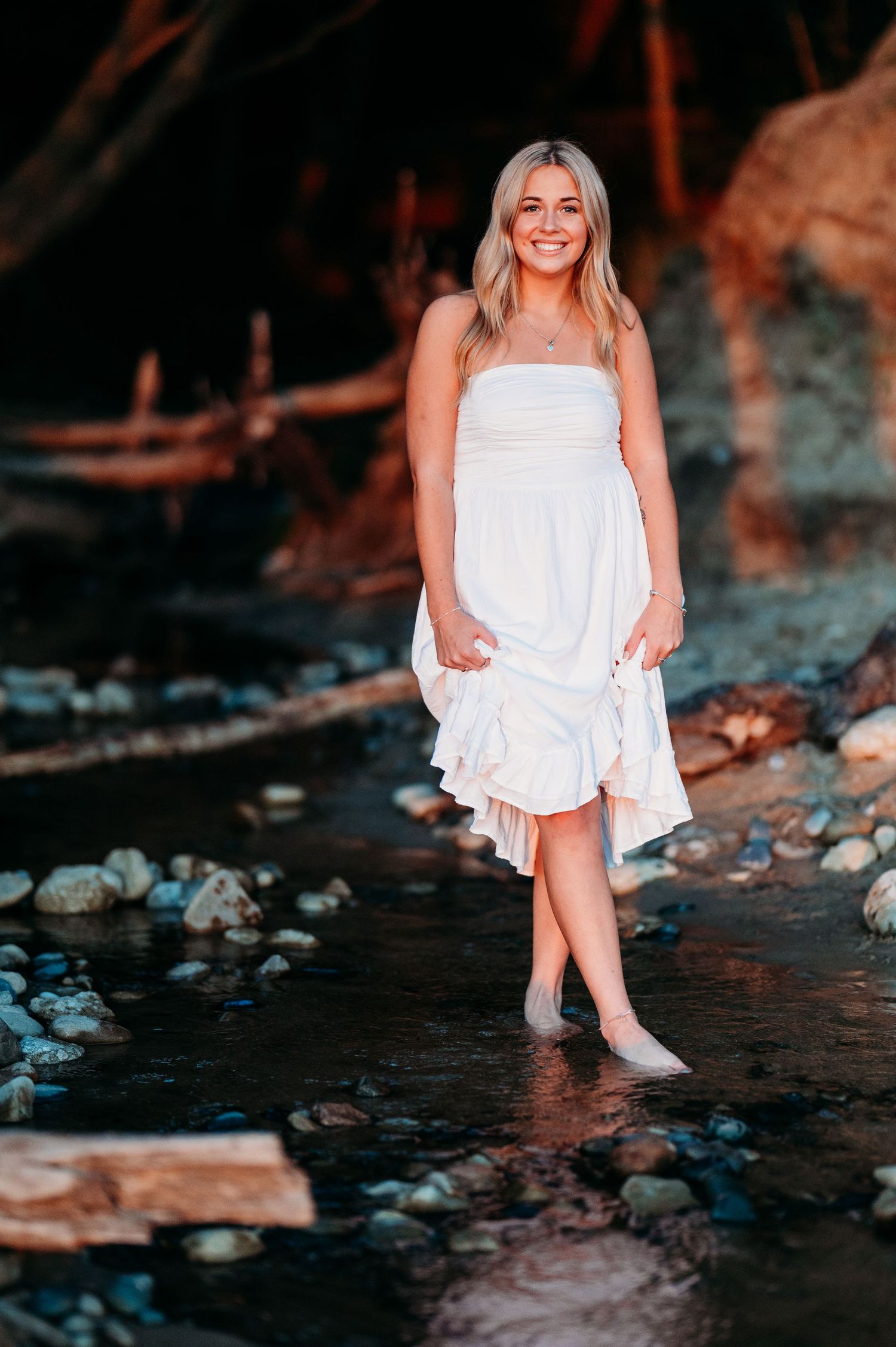 A person in a flowing white dress sits near the water's edge at sunset, creating a dreamy outdoor portrait series.