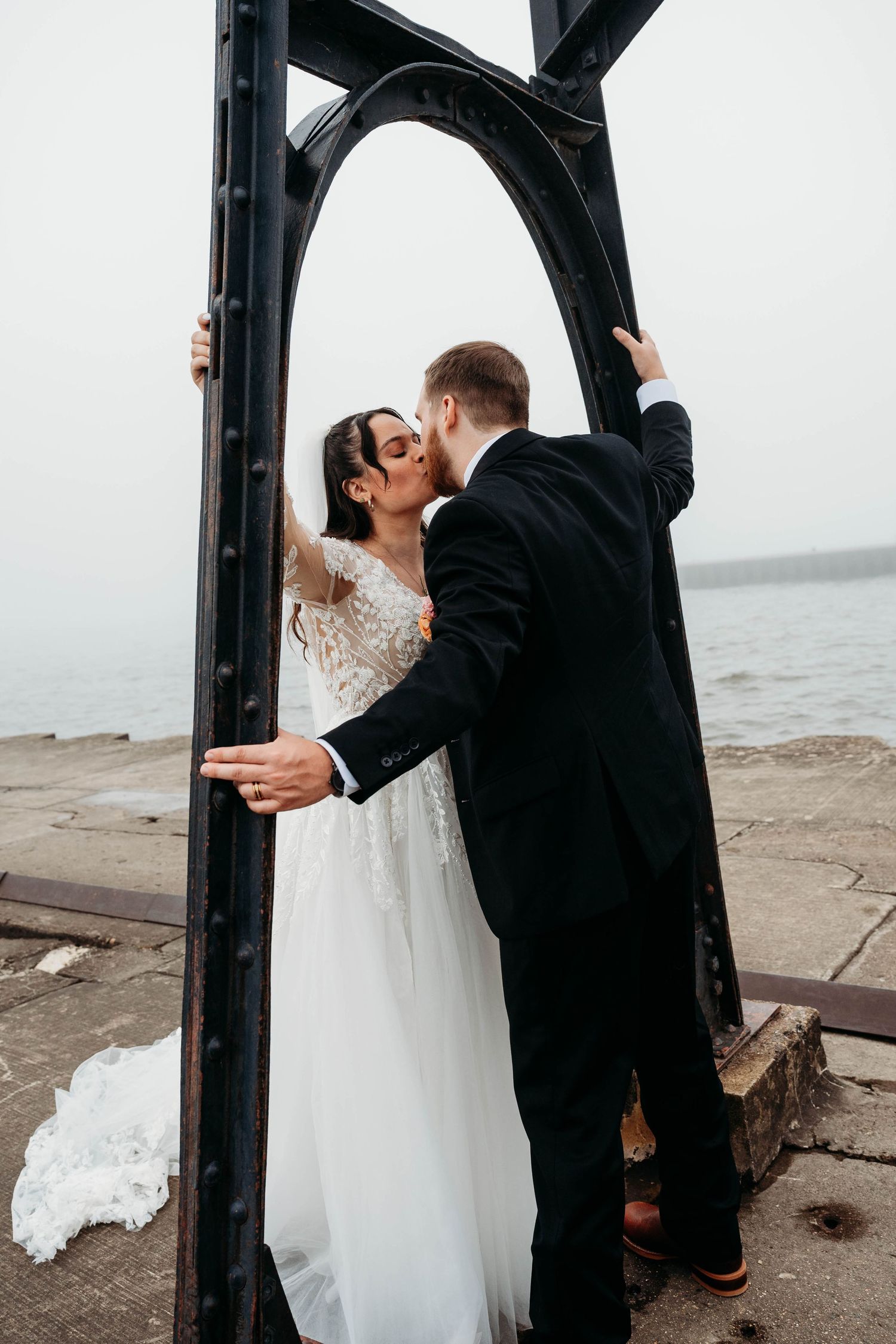 Bride and groom kiss framed by black iron arch structure beside misty waterfront on wedding day.