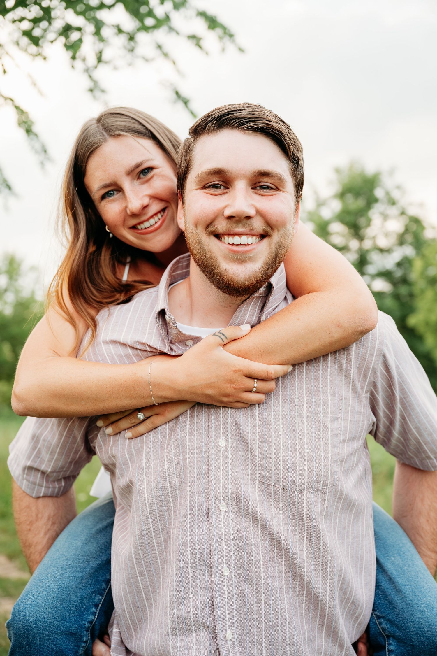 A smiling couple poses outdoors, woman giving a piggyback ride to man in striped shirt with green trees behind them.