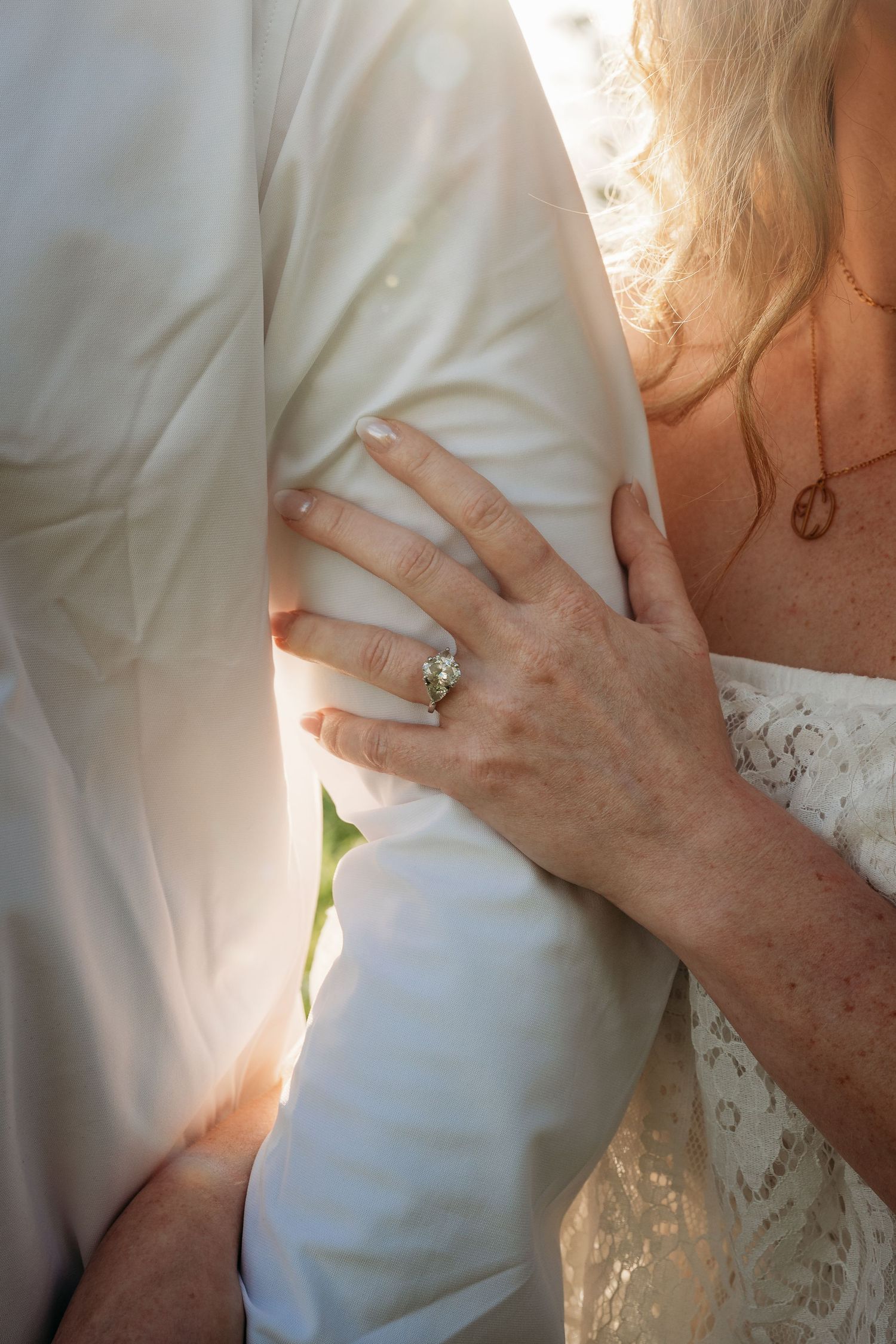 Close-up of bride's hand with diamond engagement ring resting on groom's white suit jacket.