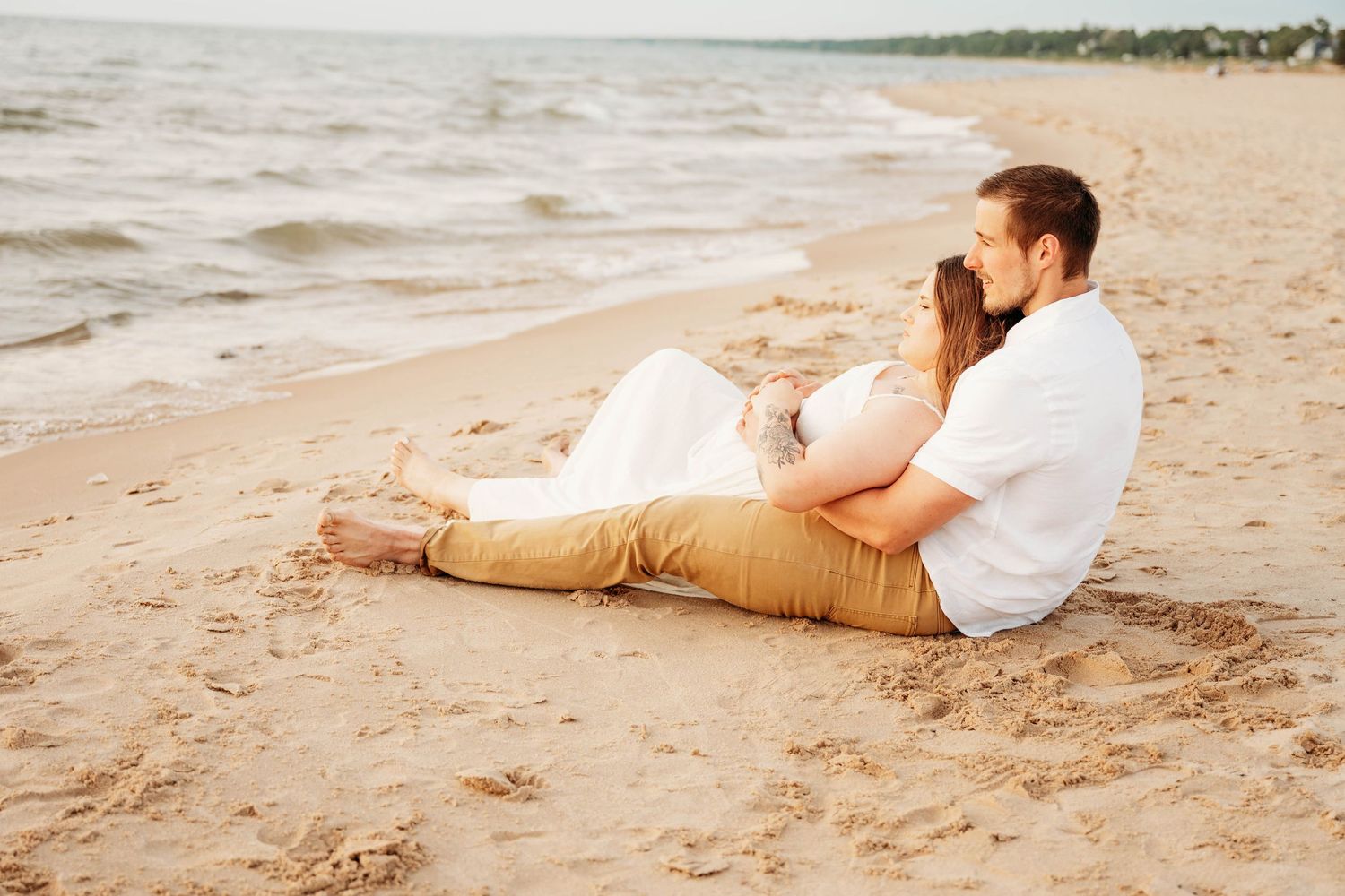 Romantic couple sitting together on sandy beach at sunset, man embracing woman in white dress near ocean waves.