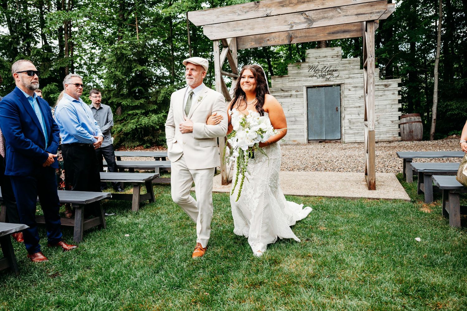 Bride walks down outdoor aisle with escort at rustic wedding venue surrounded by wooden benches and lush greenery.