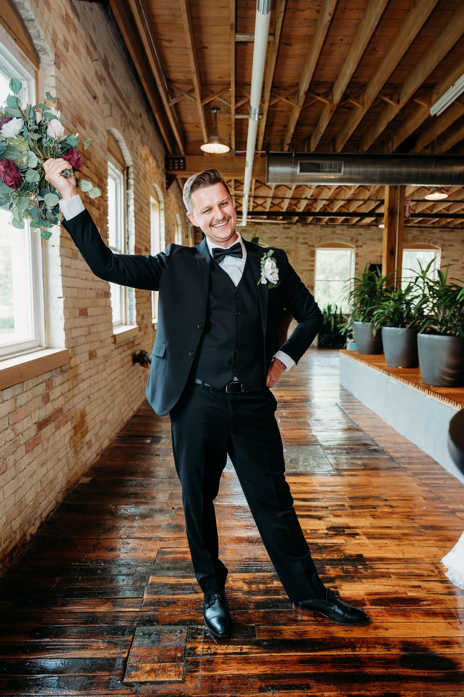 Joyful groom in black tuxedo raises bouquet triumphantly in rustic brick wedding venue with wood ceiling.