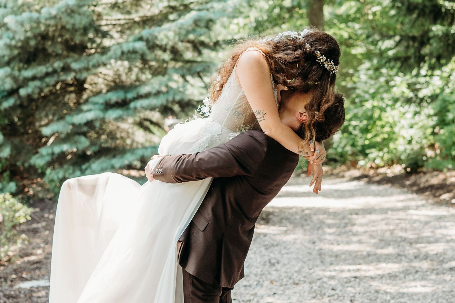 Groom dipping bride in a romantic outdoor wedding kiss surrounded by lush green trees on a gravel path.