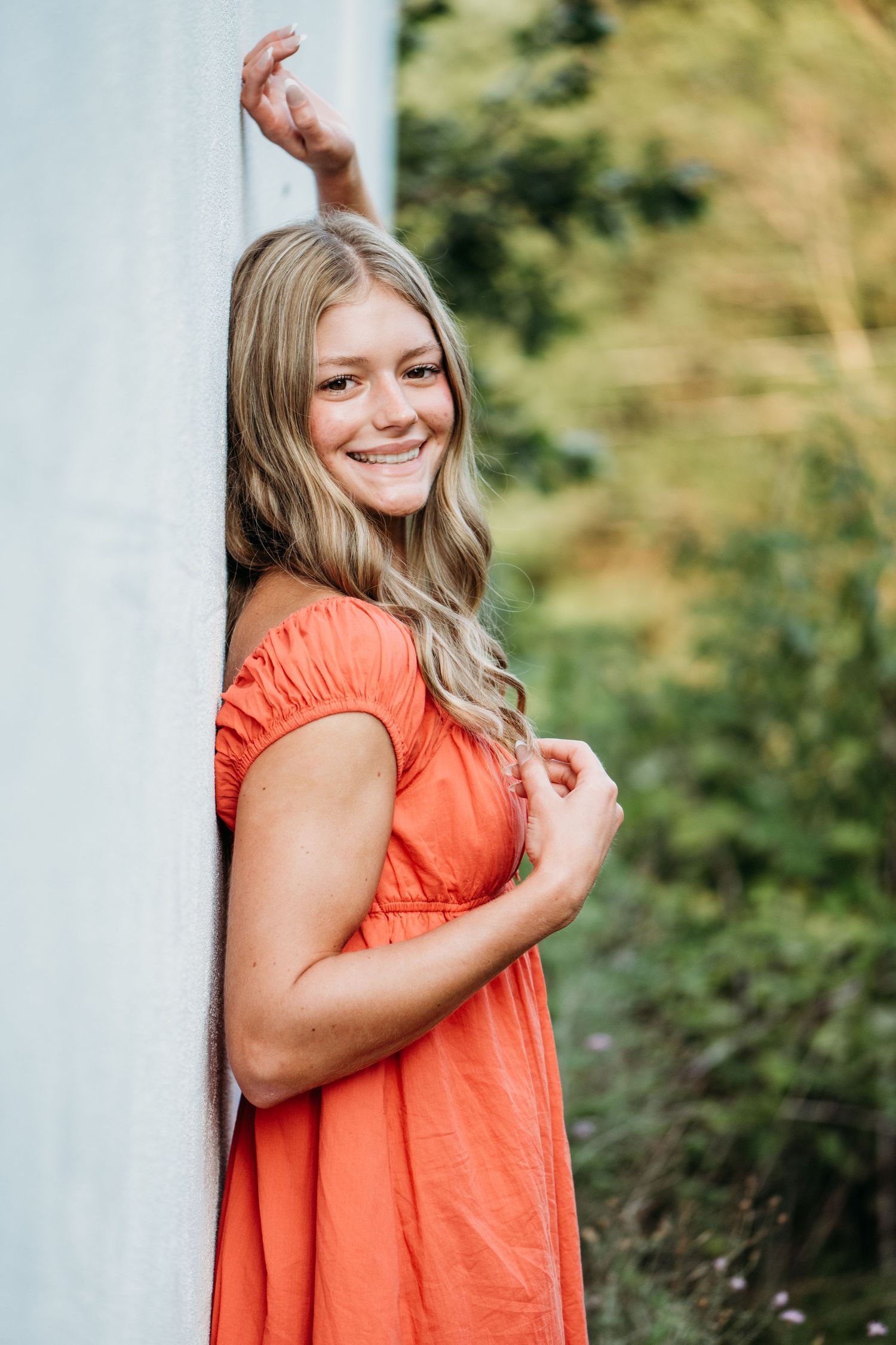 Portrait view of someone wearing a coral-colored summer dress posing against a white wall with green foliage in background.