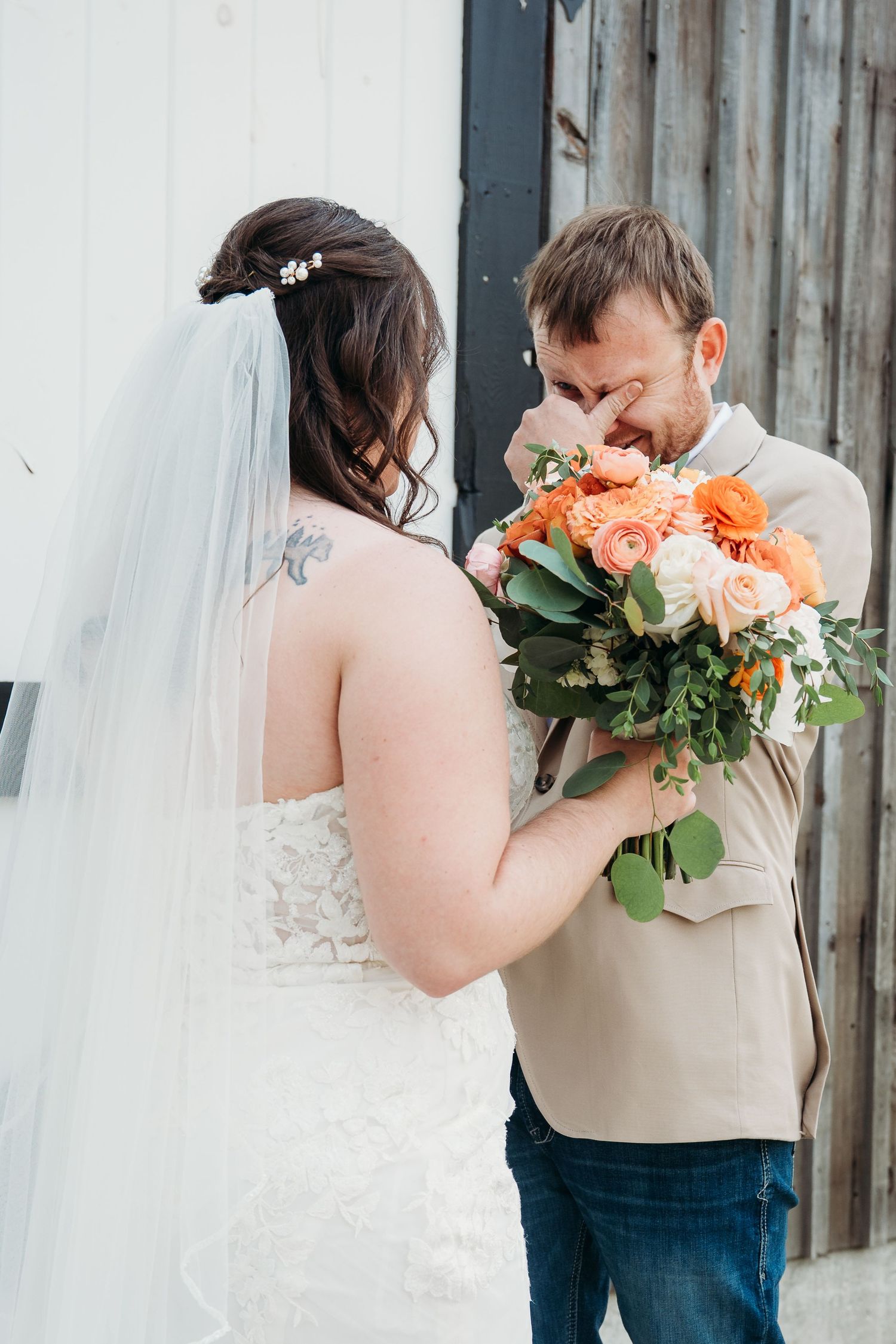 A couple shares an emotional moment with a peach and orange rose bouquet against a rustic wooden wall at their wedding.