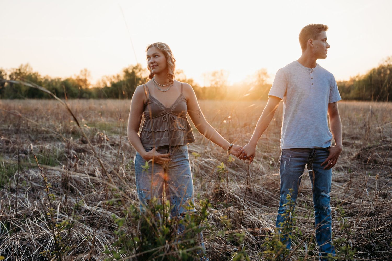 A couple holds hands while walking through a golden sunlit field during sunset, creating a romantic silhouette.