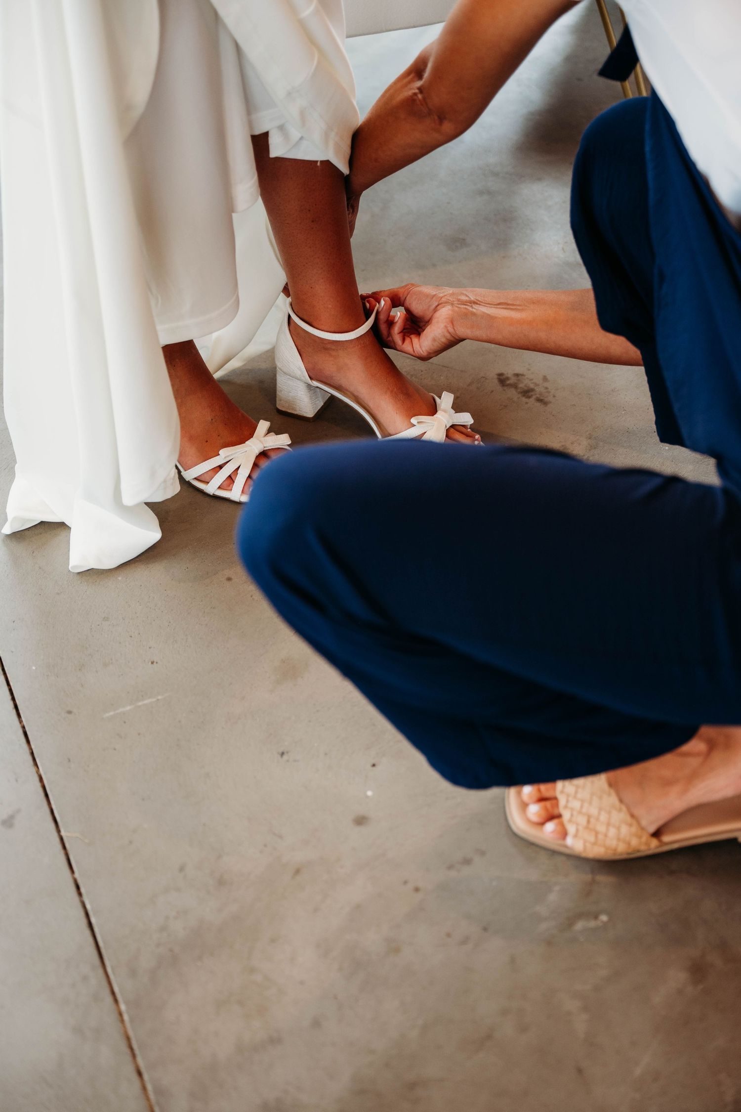 Close-up shot of someone helping adjust white strappy sandals on a bride's feet during wedding preparations.