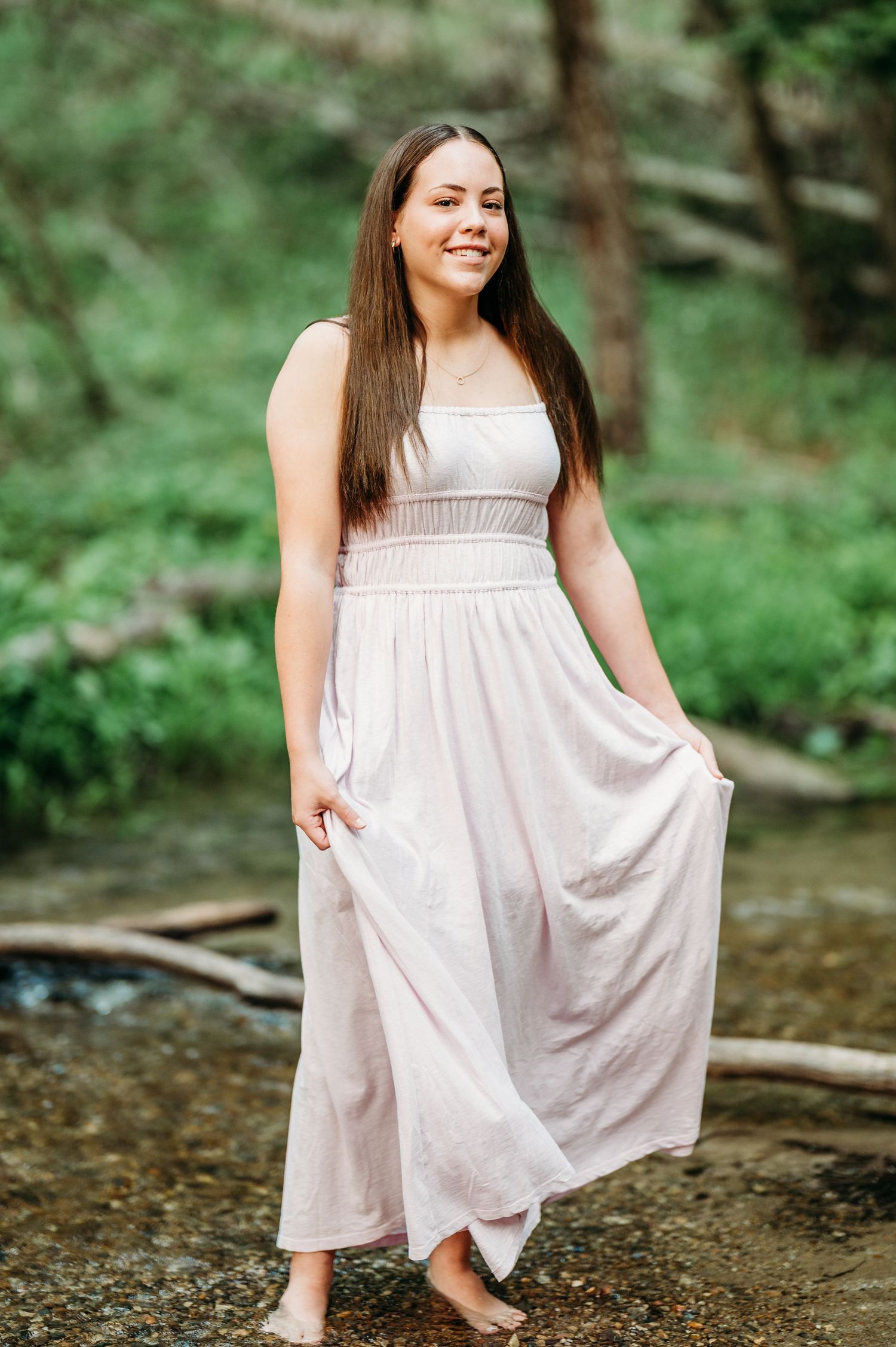 White flowing sundress twirls gracefully by a creek in a lush forest setting on a sunny afternoon.
