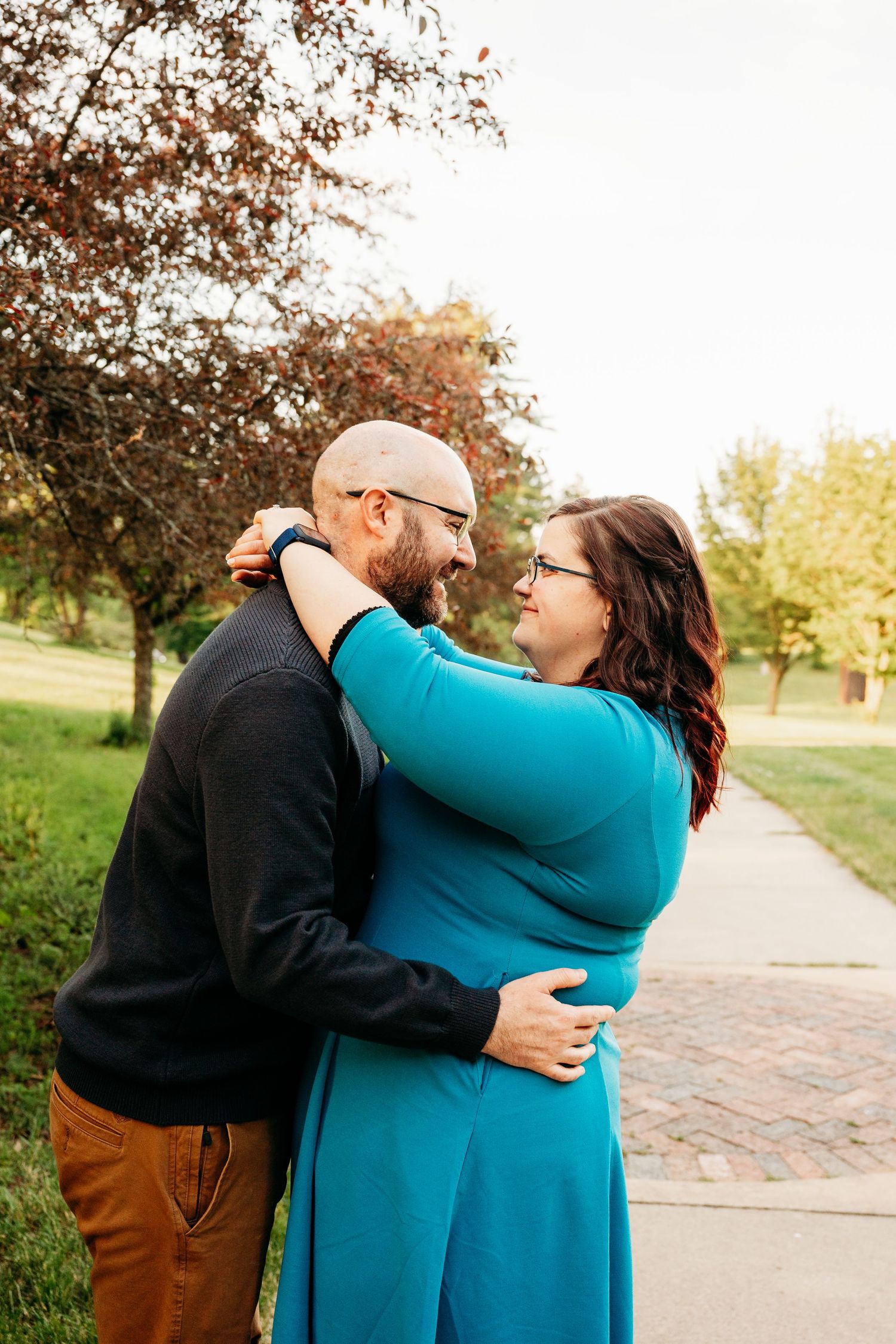 A couple embraces and smiles at each other on a park path, she wears a teal dress and he wears a black sweater.