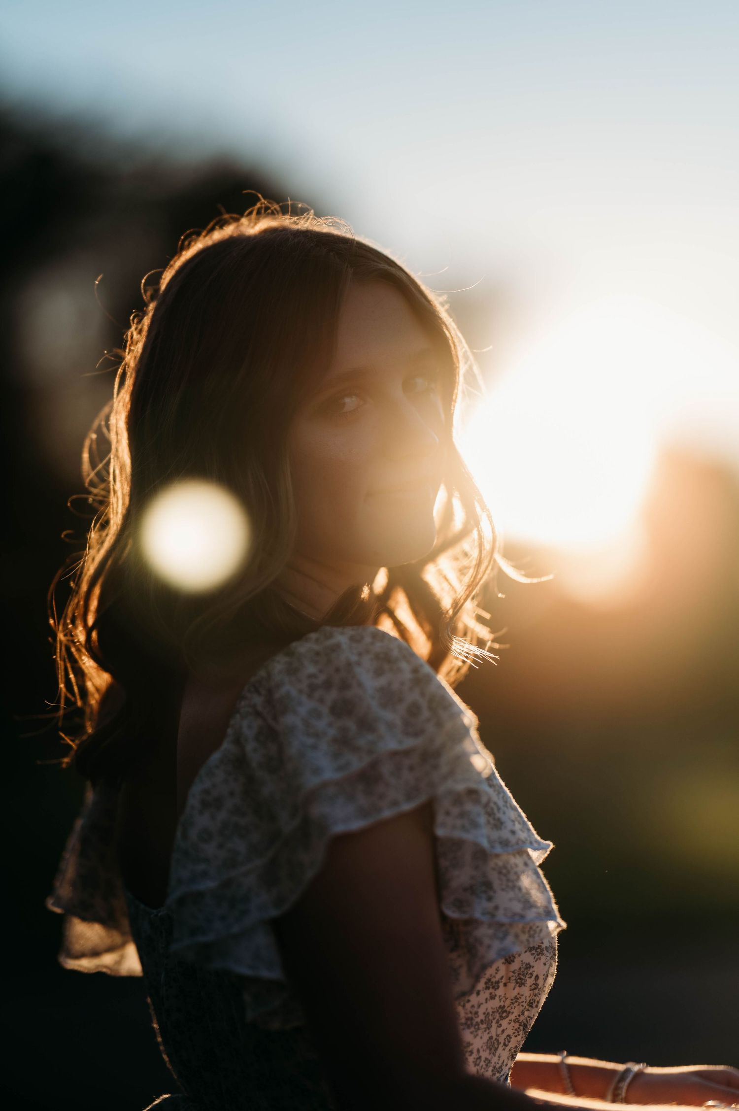 A young girl with flowing hair bathed in golden sunset backlight wearing a floral off-shoulder top outdoors.