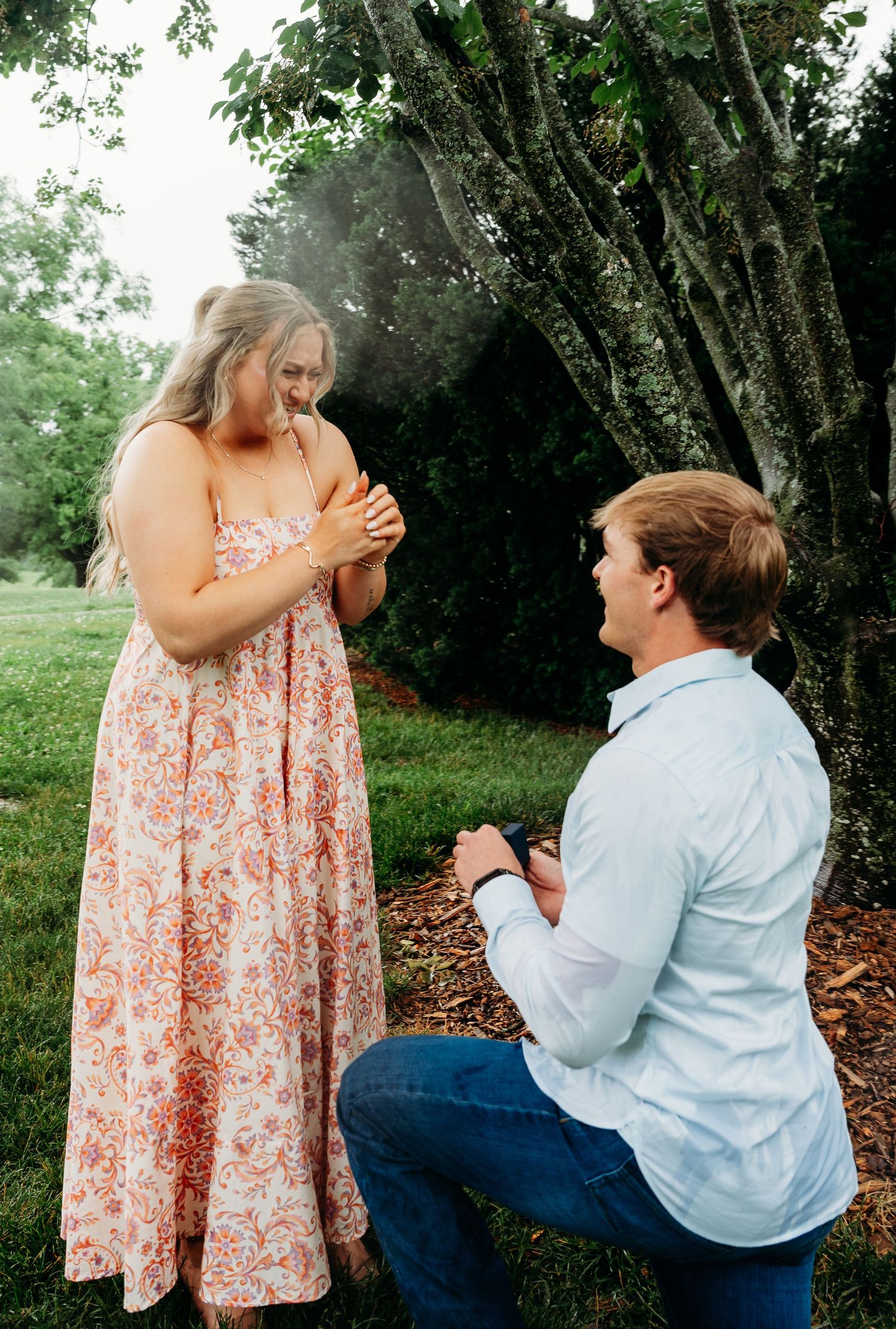 Man kneeling on one knee proposing to a woman in a floral dress outdoors in a lush green garden.