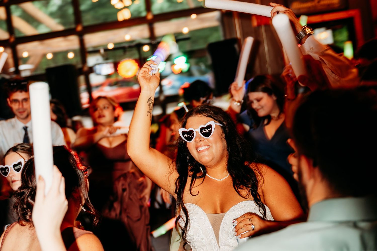 Bride dancing joyfully in heart-shaped sunglasses at a lively wedding reception party with guests celebrating.
