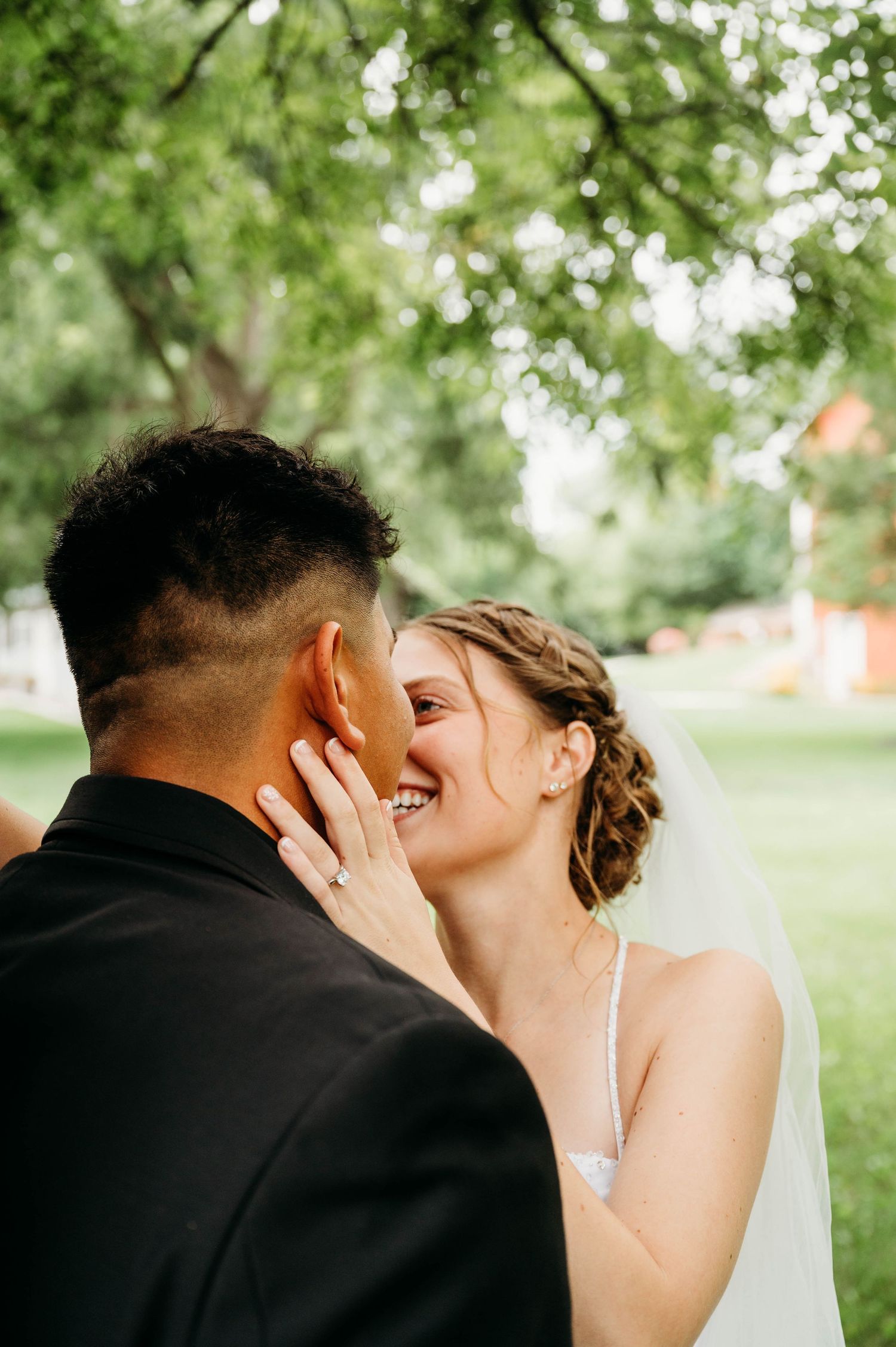 Smiling bride in white dress and veil embraces groom in black suit outdoors under green trees on wedding day.