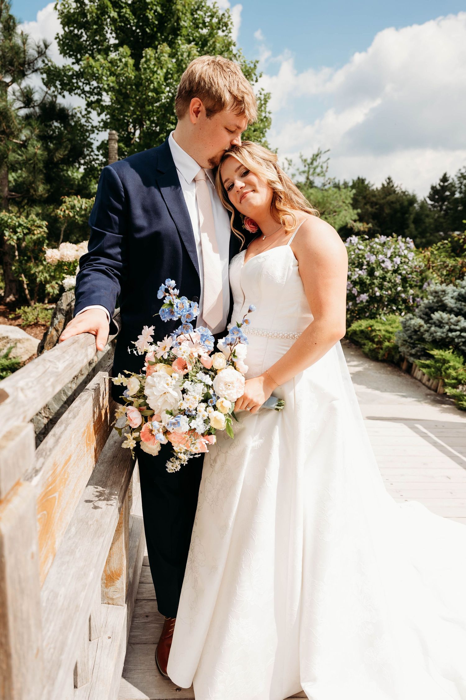 Bride and groom share a tender moment on a garden bridge, she holds a colorful bouquet of pink, white, and blue flowers.