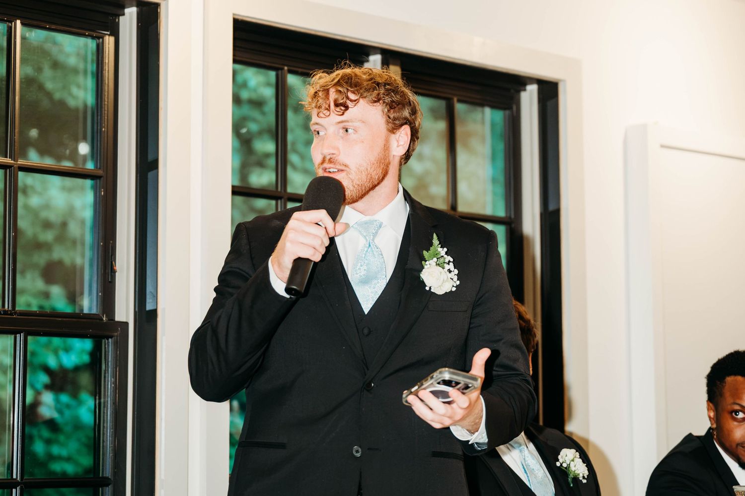 Groom in black suit with boutonniere giving wedding speech holding microphone at elegant indoor reception.