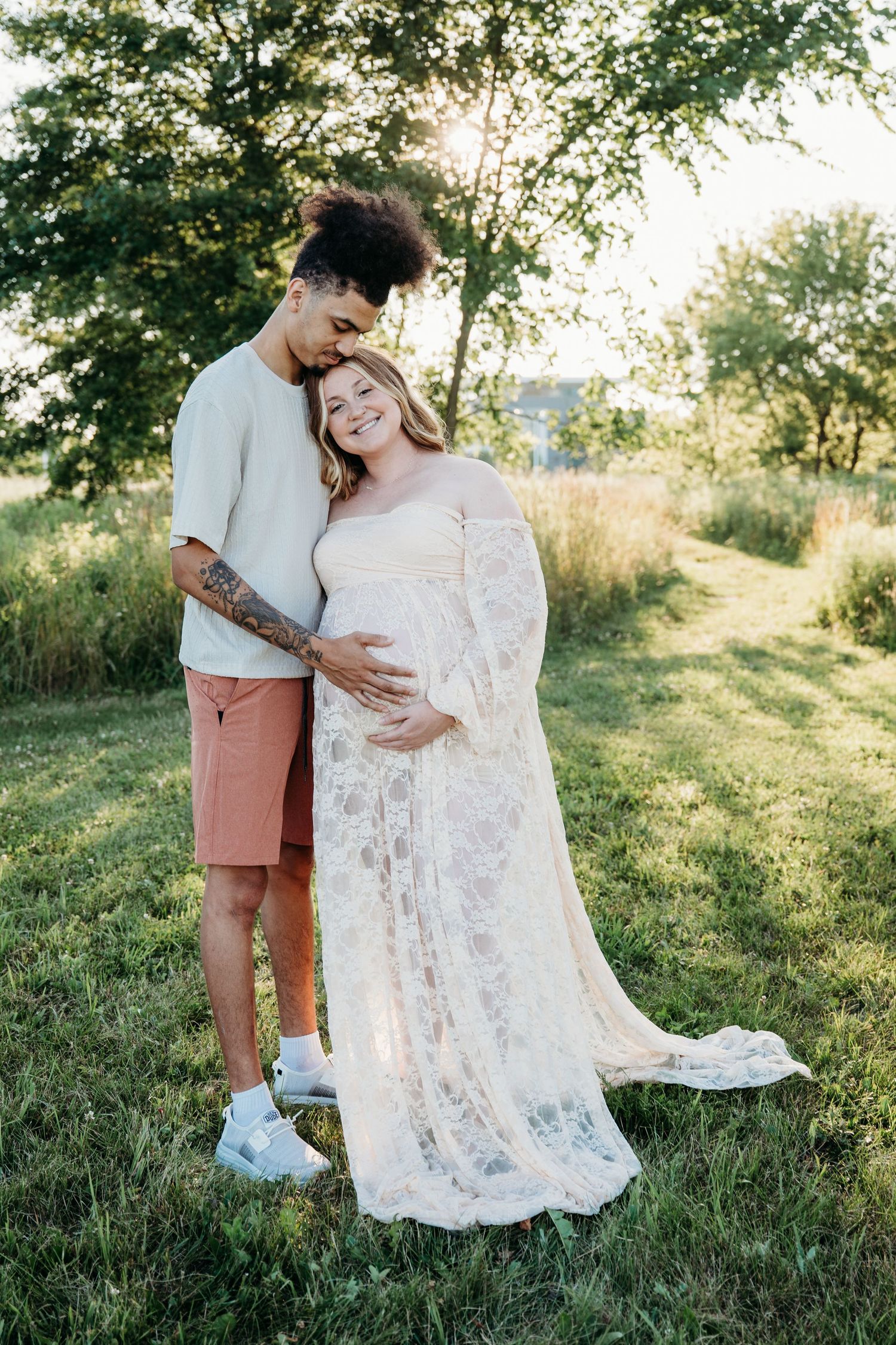 A couple embraces in a sunlit field, with her wearing a flowing lace dress and him in casual summer attire.