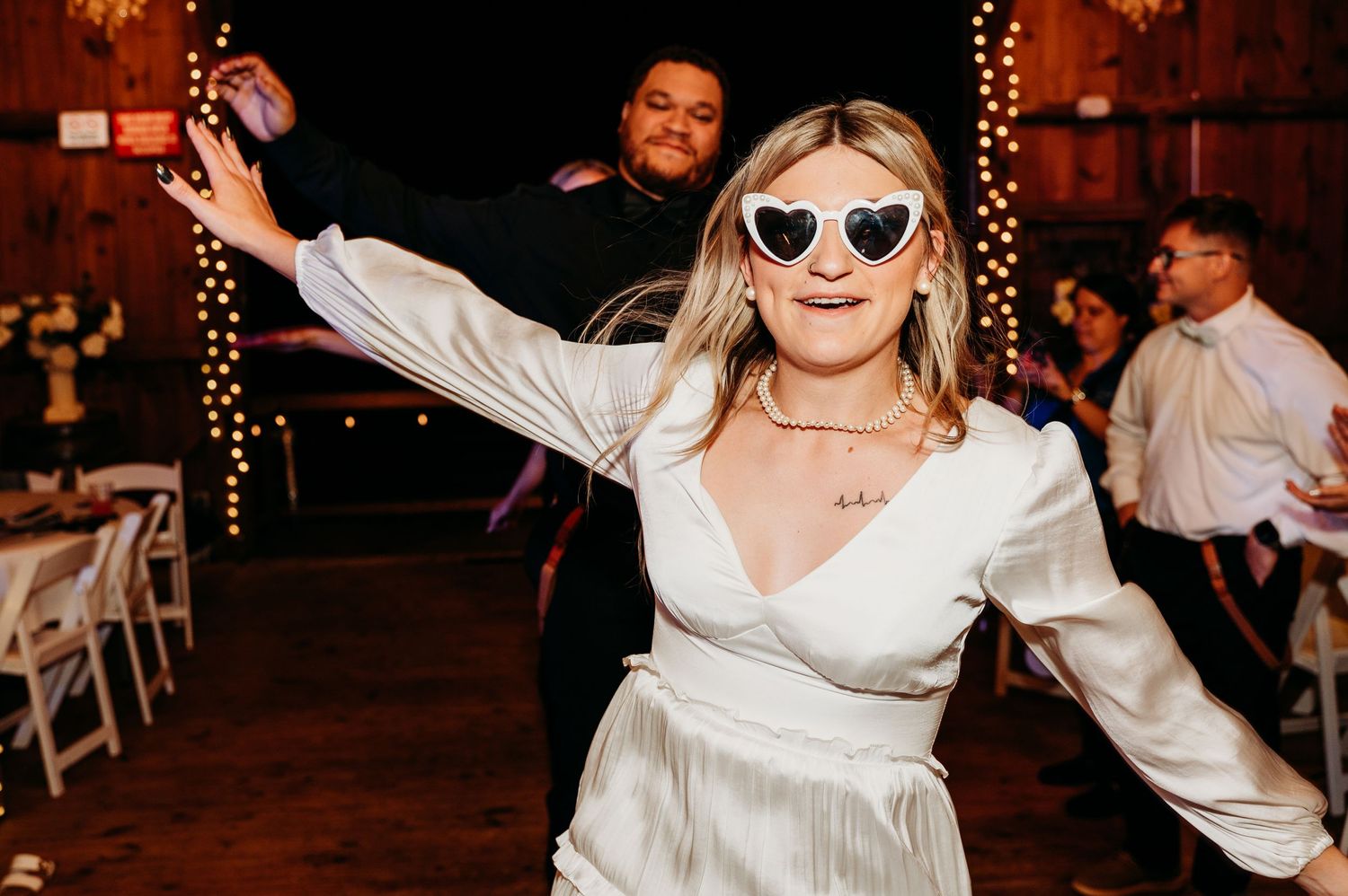 Person in white dress and sunglasses dances joyfully at an evening reception with string lights in background.