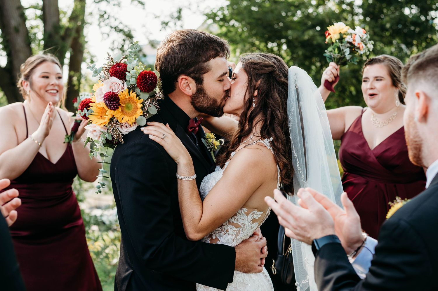 Bride and groom share first kiss surrounded by bridesmaids in burgundy dresses holding colorful bouquets at outdoor wedding.