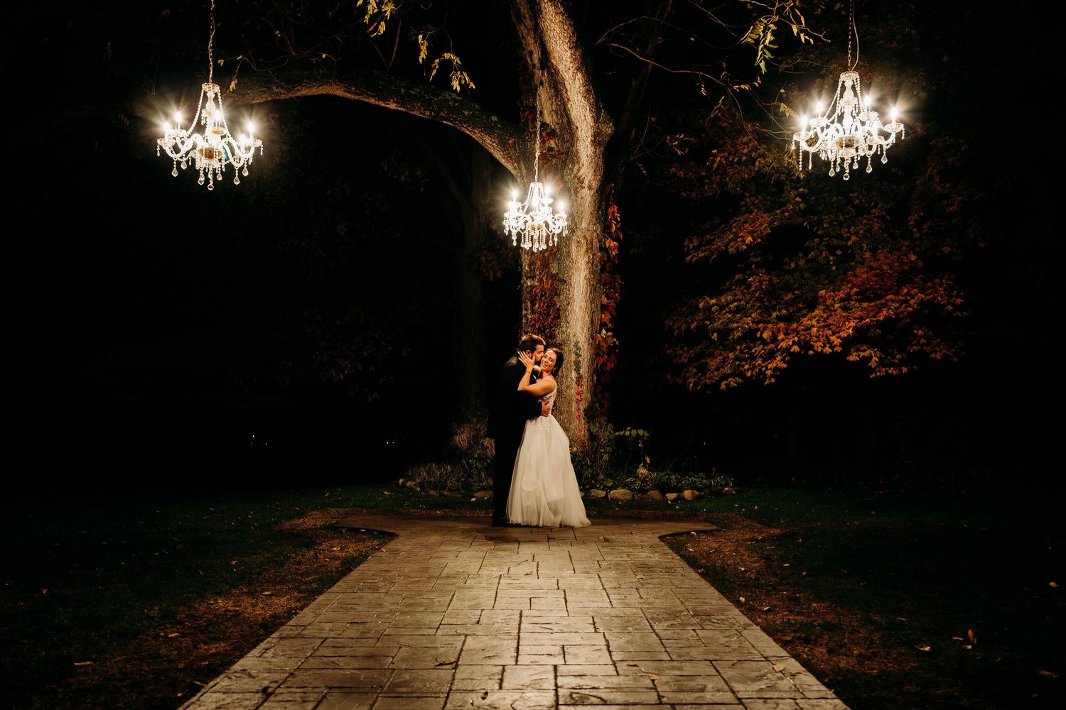 A romantic nighttime wedding scene with illuminated chandeliers and a stone path through a garden.