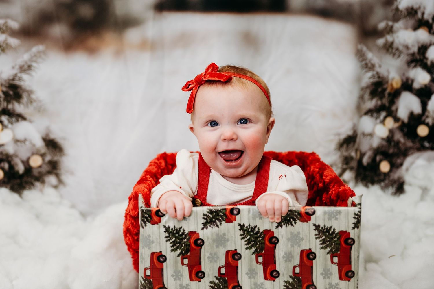 Baby in a red dress sitting in a wooden crate surrounded by Christmas decorations.
