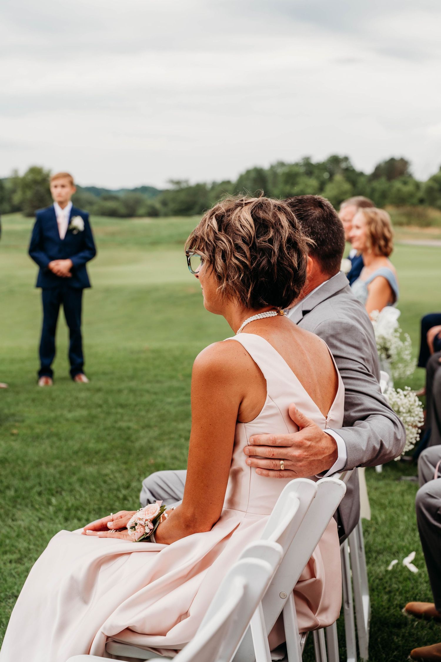 Wedding guests seated in white chairs on a grassy lawn during an outdoor ceremony.