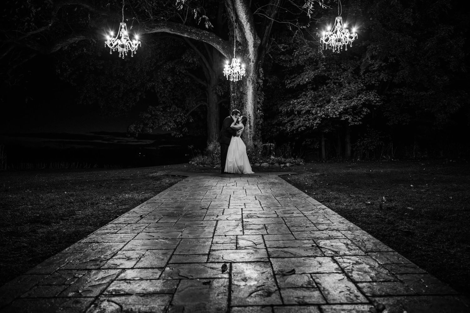 Black and white wedding photo on stone path with hanging crystal chandeliers and dramatic lighting.