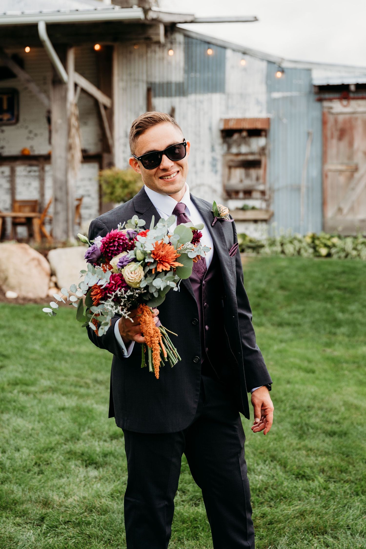 Wedding party member in black suit and sunglasses holding colorful autumn bouquet on grassy lawn by rustic buildings.