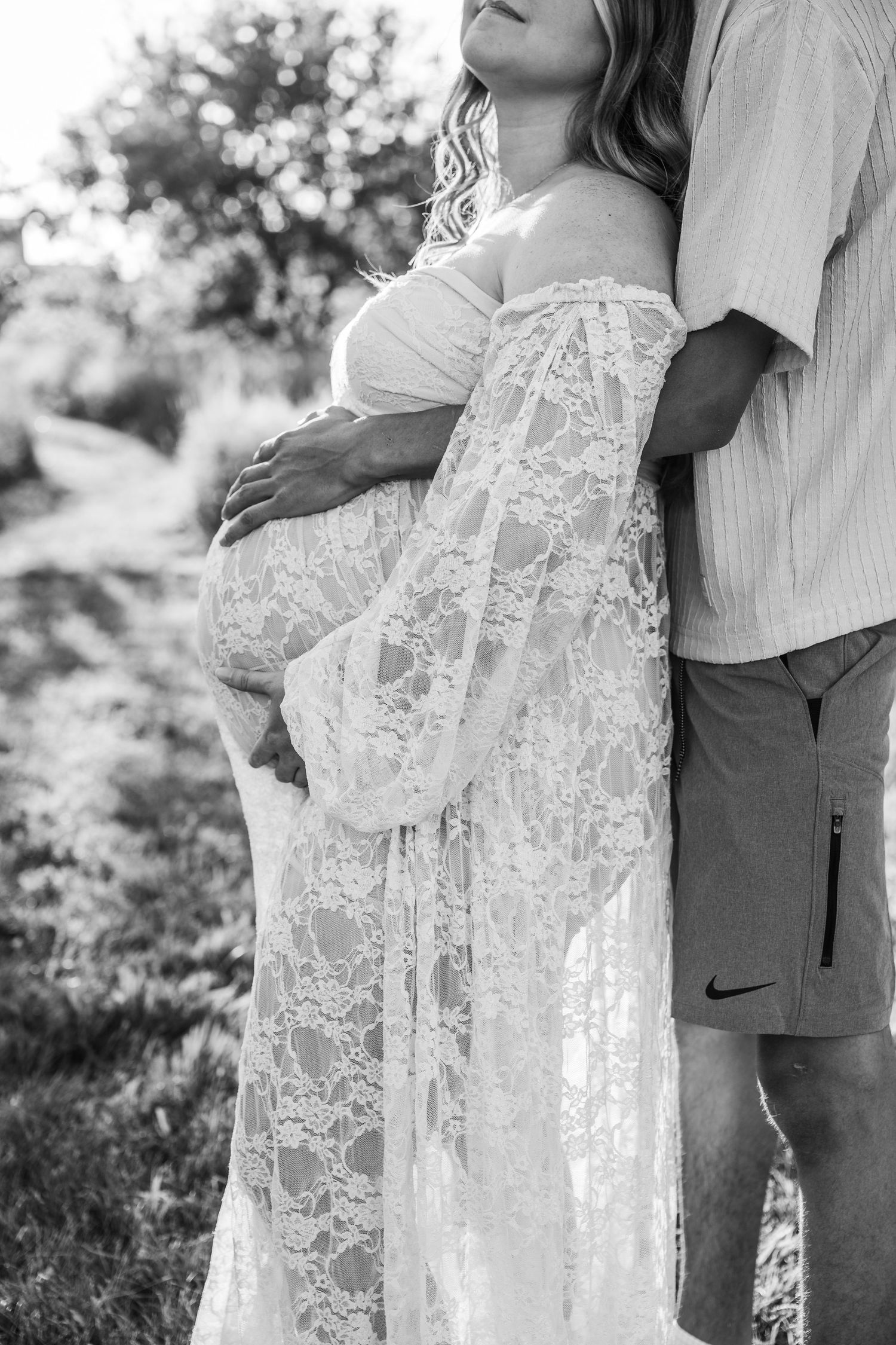 A black and white maternity photoshoot showing close-up details of a delicate white lace dress against casual attire.