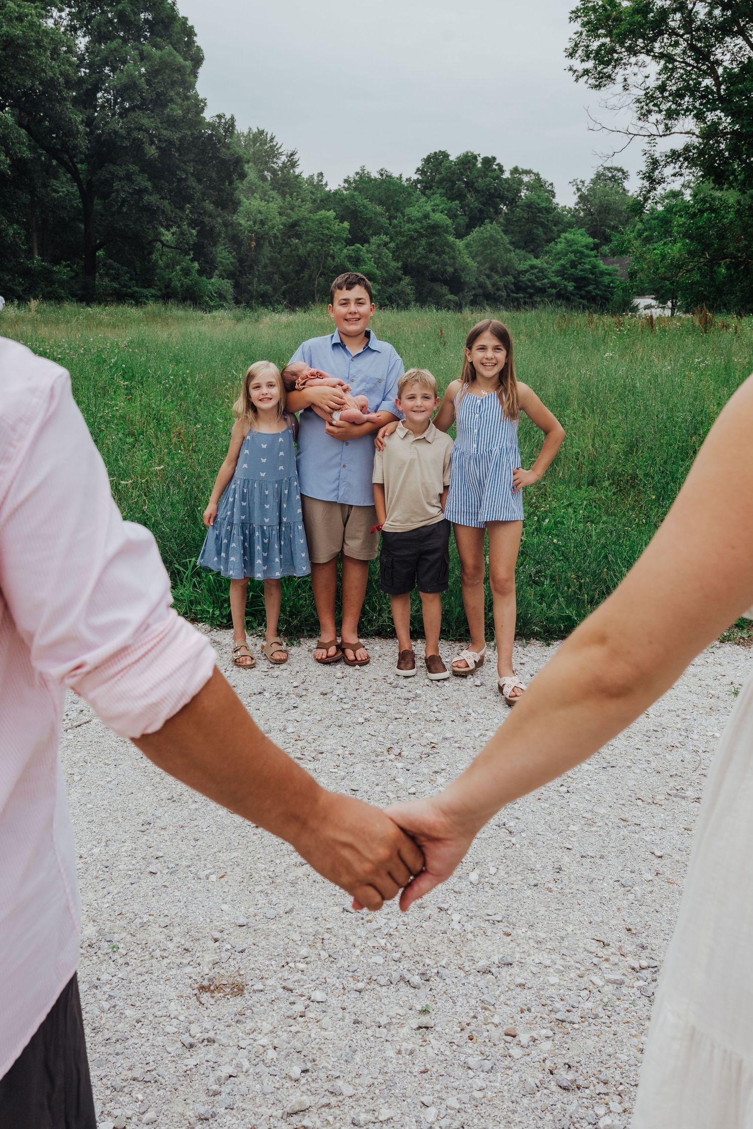 A couple holding hands in the foreground while a family stands on a gravel path surrounded by green meadow.