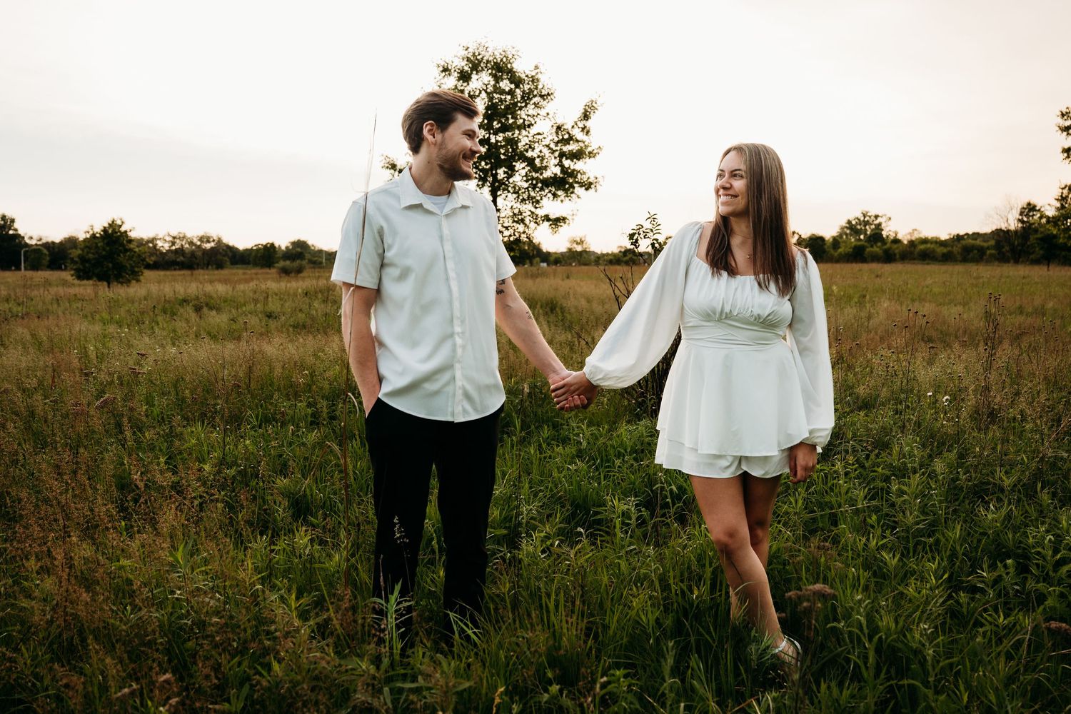 A couple shares intimate moments while walking through a golden sunset field wearing white and light blue clothing.