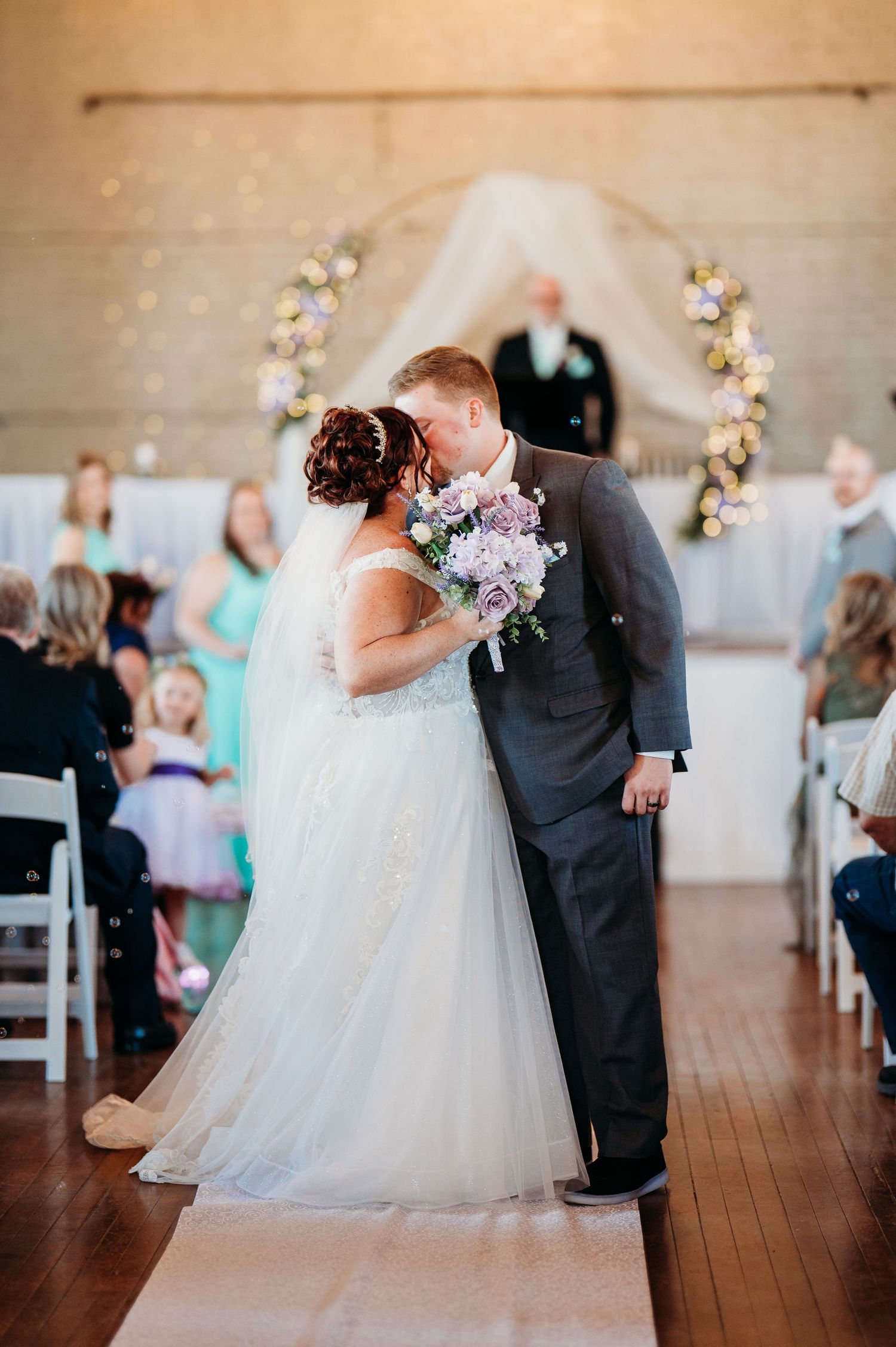 A tender wedding ceremony moment captured in a bright indoor venue with floral arch and wooden floors.