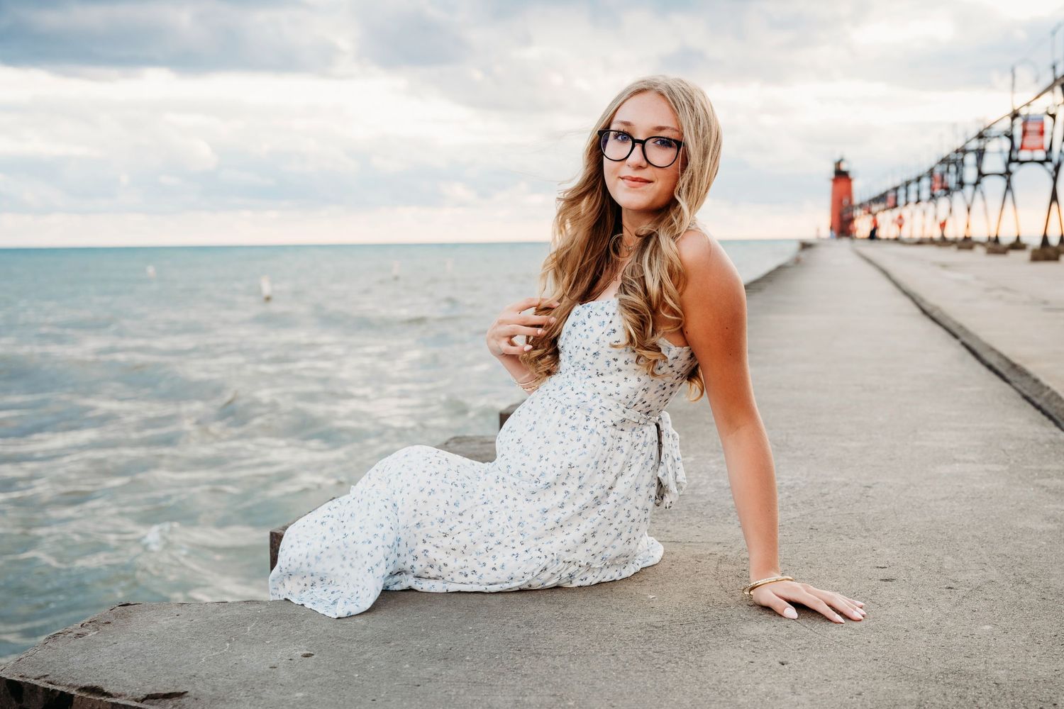 A young woman in a white dress and glasses poses on a pier by the ocean during a sunny afternoon.