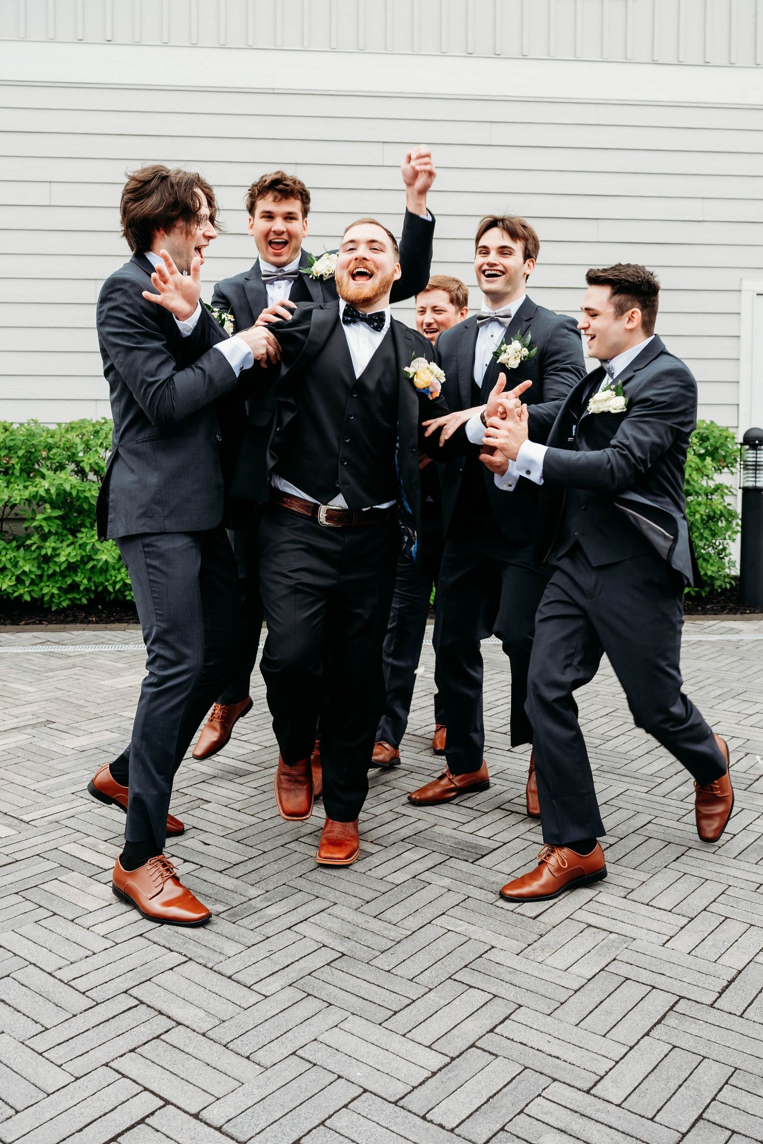Groom and groomsmen in black tuxedos with brown shoes celebrating joyfully on a brick patio.