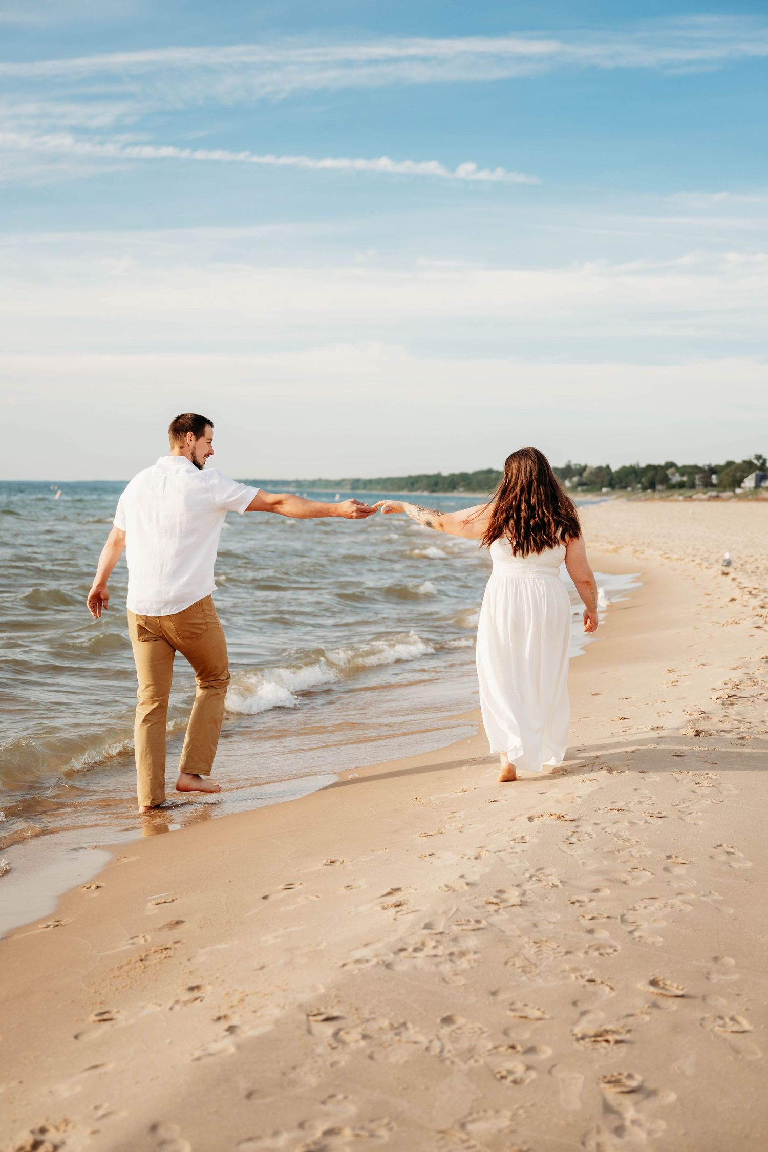Couple holding hands walking barefoot along sandy beach shoreline, woman in white dress and man in white shirt and tan pants.