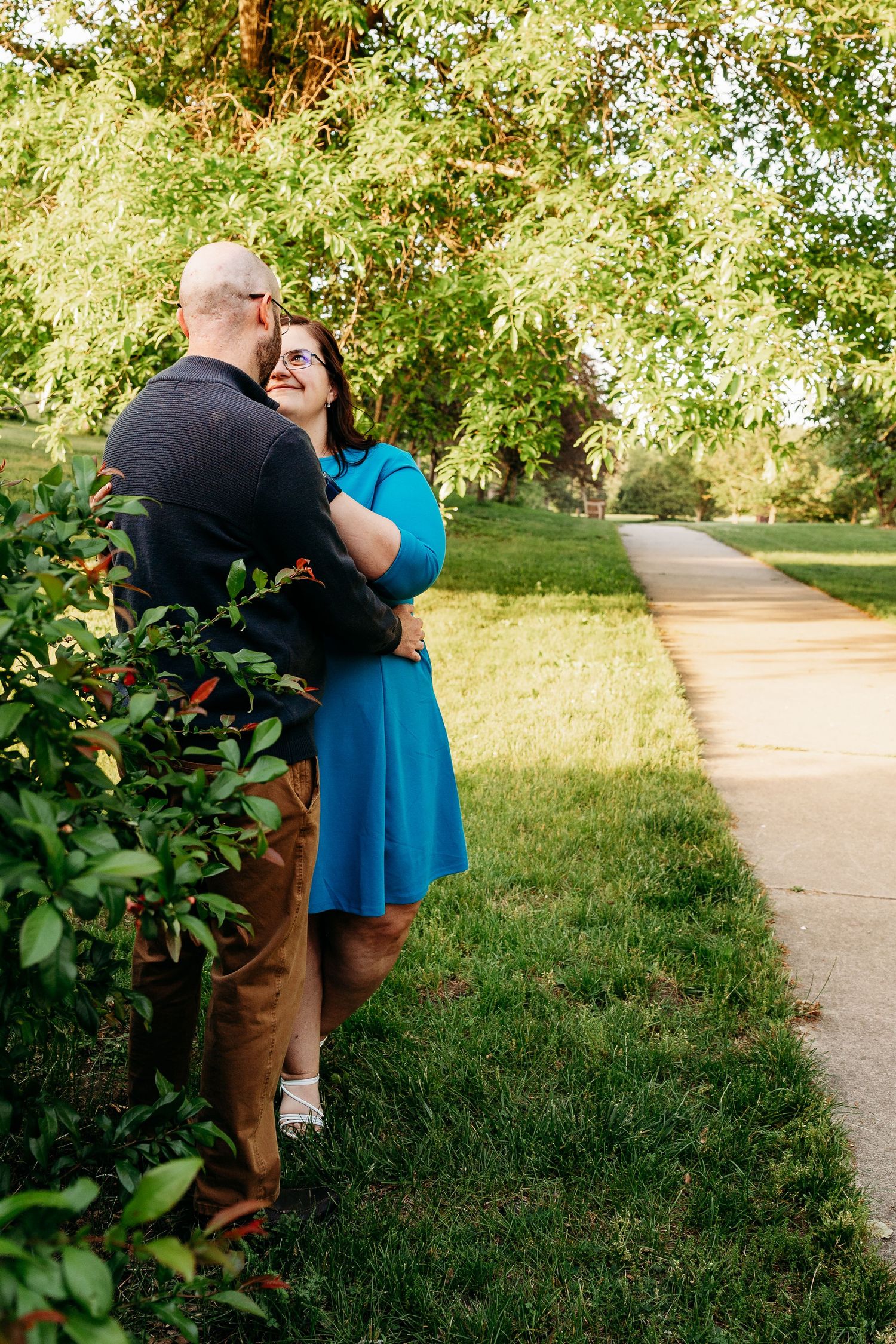 Couple embracing and smiling near lush green bushes along a sunny park pathway, woman in blue dress.